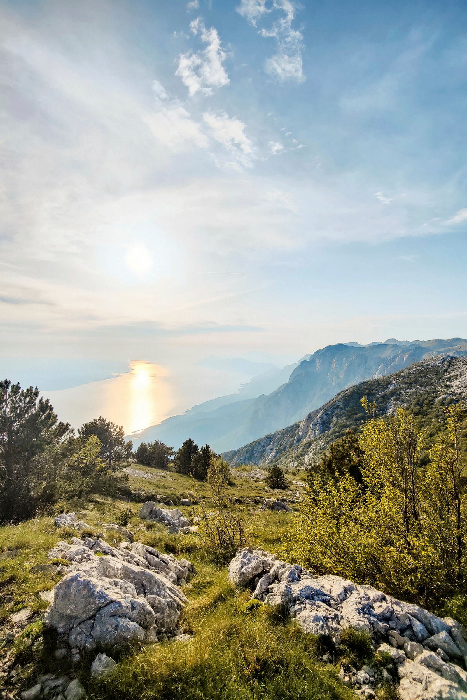 Utsikt over et fjellandskap med vegetasjon, steiner og trær, og havet i bakgrunnen ved solnedgang. (AI-generert tekst)