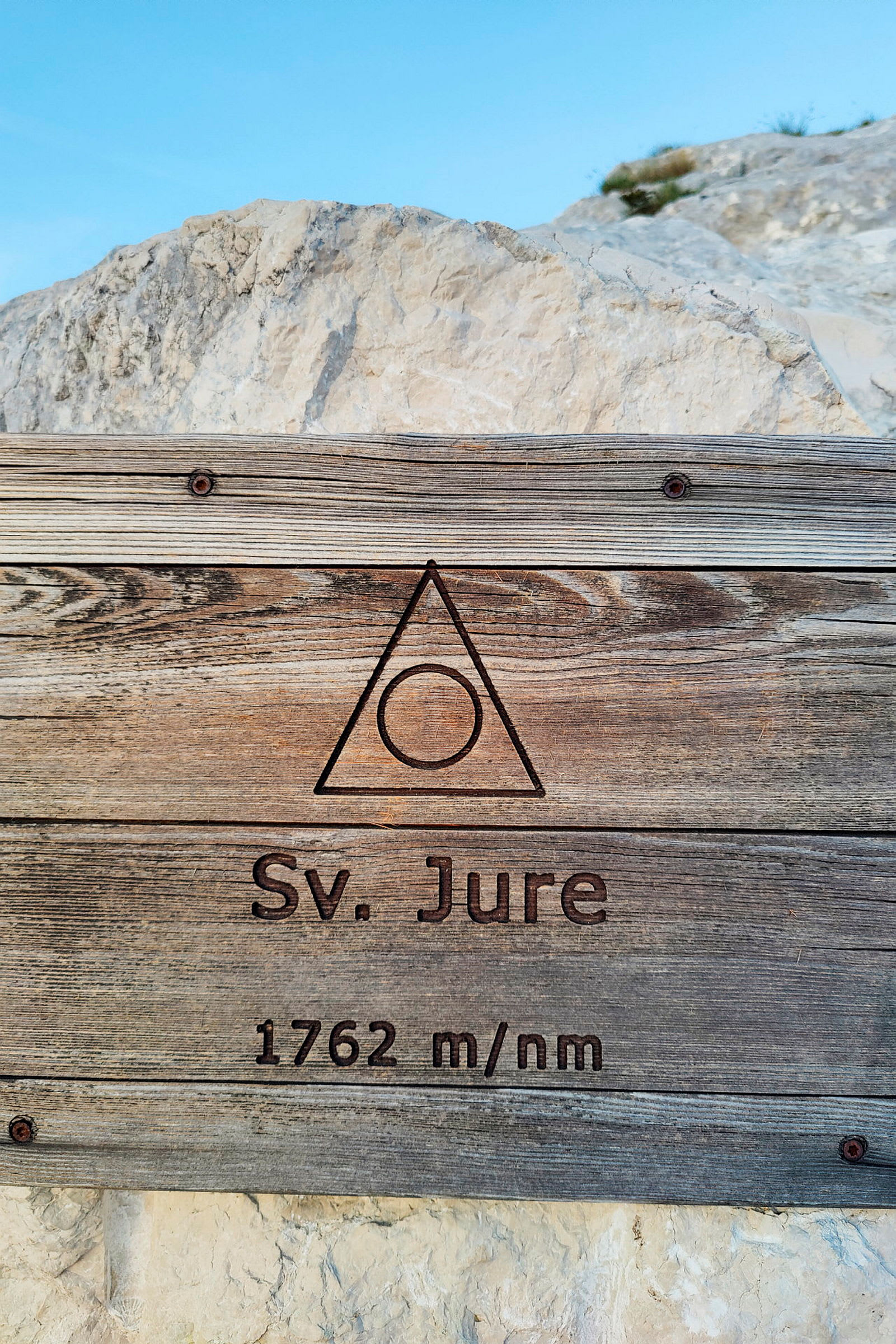Treskilt med teksten 'Sv. Jure 1762 m/nm' mot en bakgrunn av klipper og blå himmel. (AI-generert tekst)