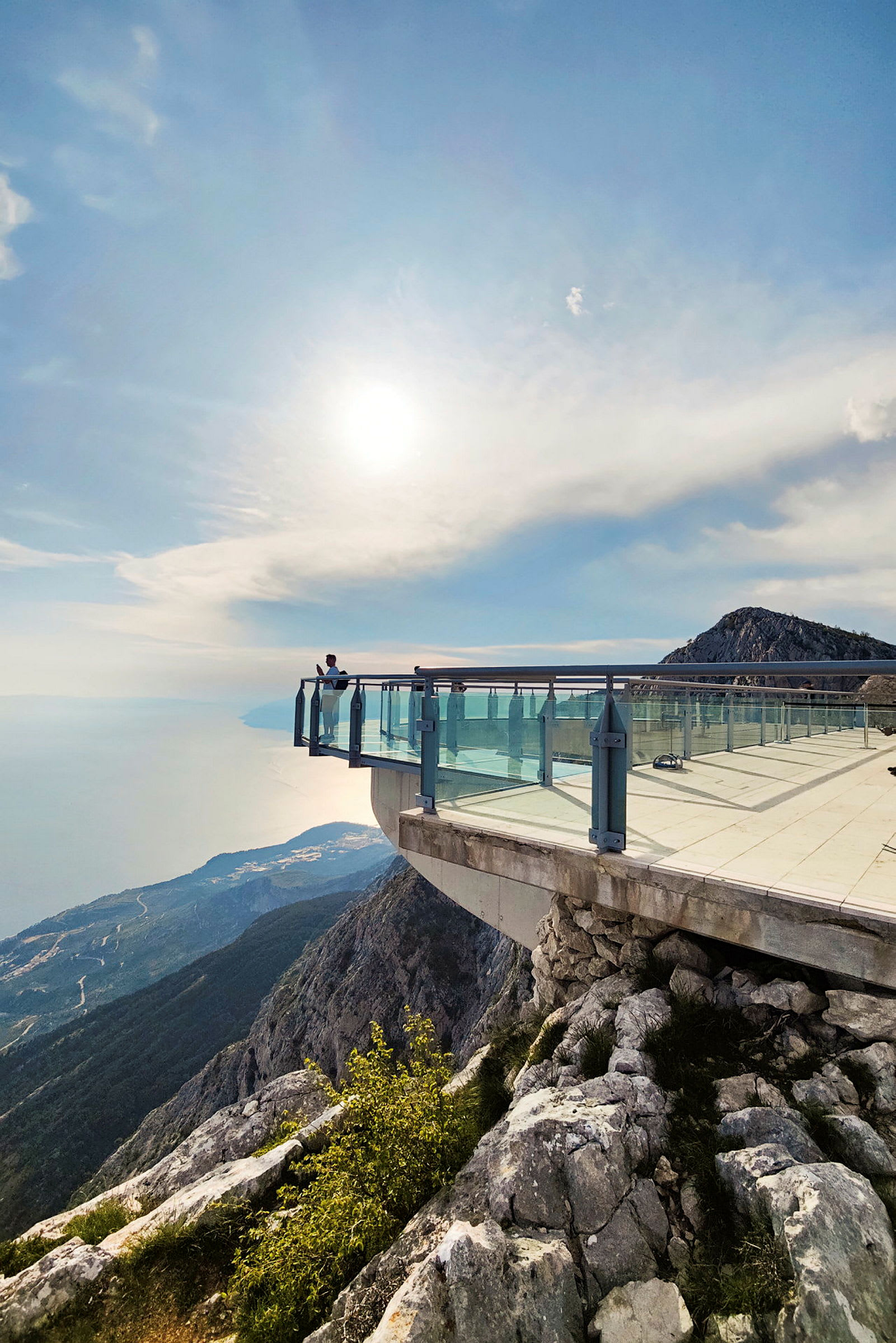En utsiktsplattform med glassrekkverk som strekker seg ut over en klippe. Solen skinner og gir et mykt lys over fjellandskapet og havet nedenfor. (AI-generert tekst)