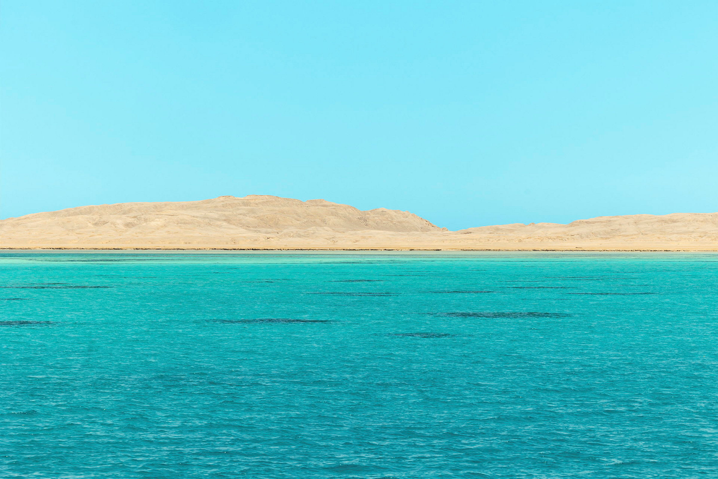 En klar blå himmel over et turkis hav med en sandstrand og åser i bakgrunnen. (AI-generert tekst)