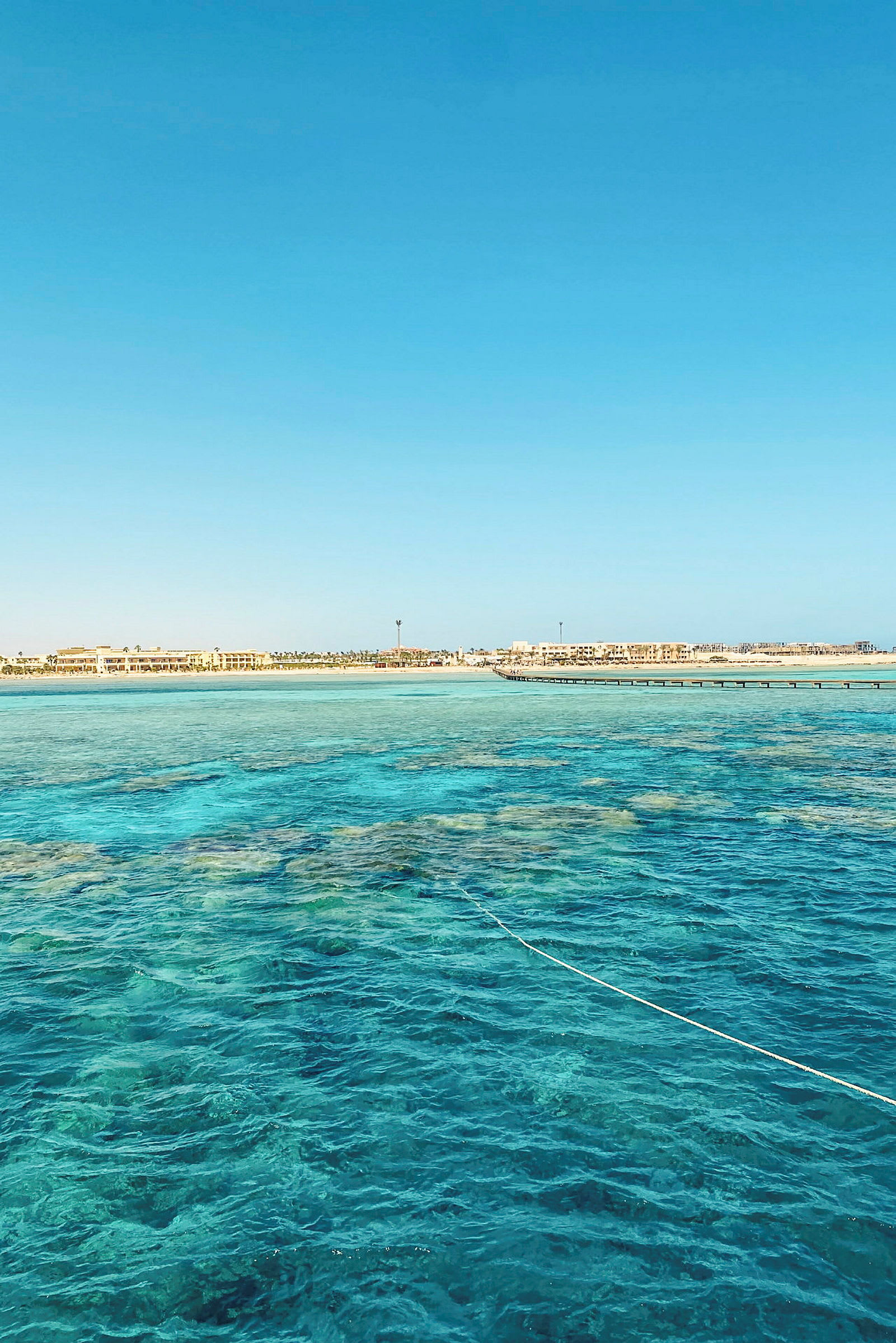 Bilde av et klart blått hav med korallstrukturer under overflaten og en linje som strekker seg over vannet. Strandlinje og bygninger vises i bakgrunnen. (AI-generert tekst)