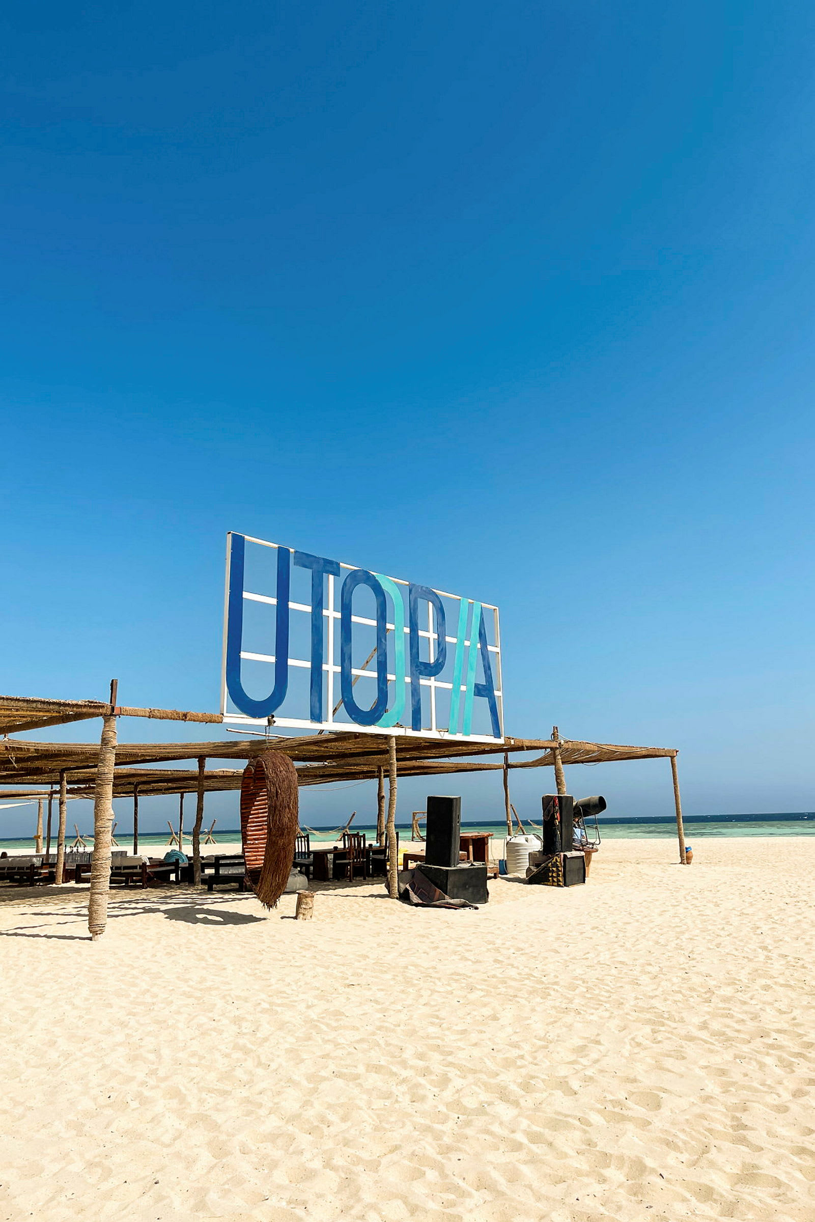 Skilt med teksten 'UTOPIA' på en strand med sitteplasser og rekvisita. (AI-generert tekst)