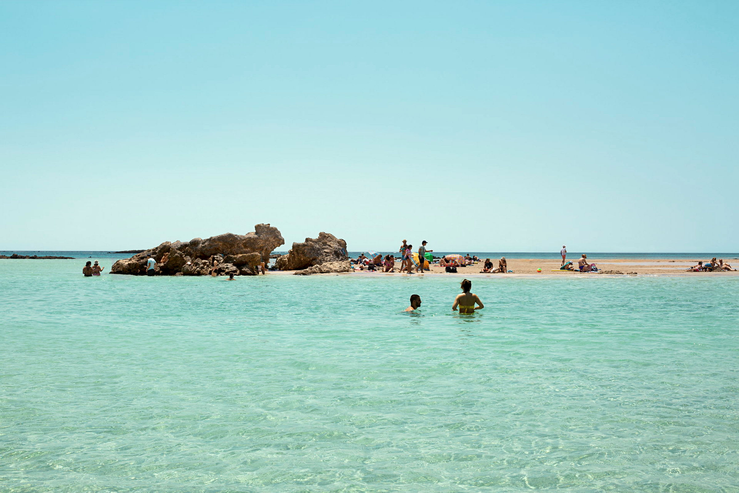 En strand med steiner og krystallklart vann. Folk nyter strandlivet og svømmer i havet. (AI-generert tekst)