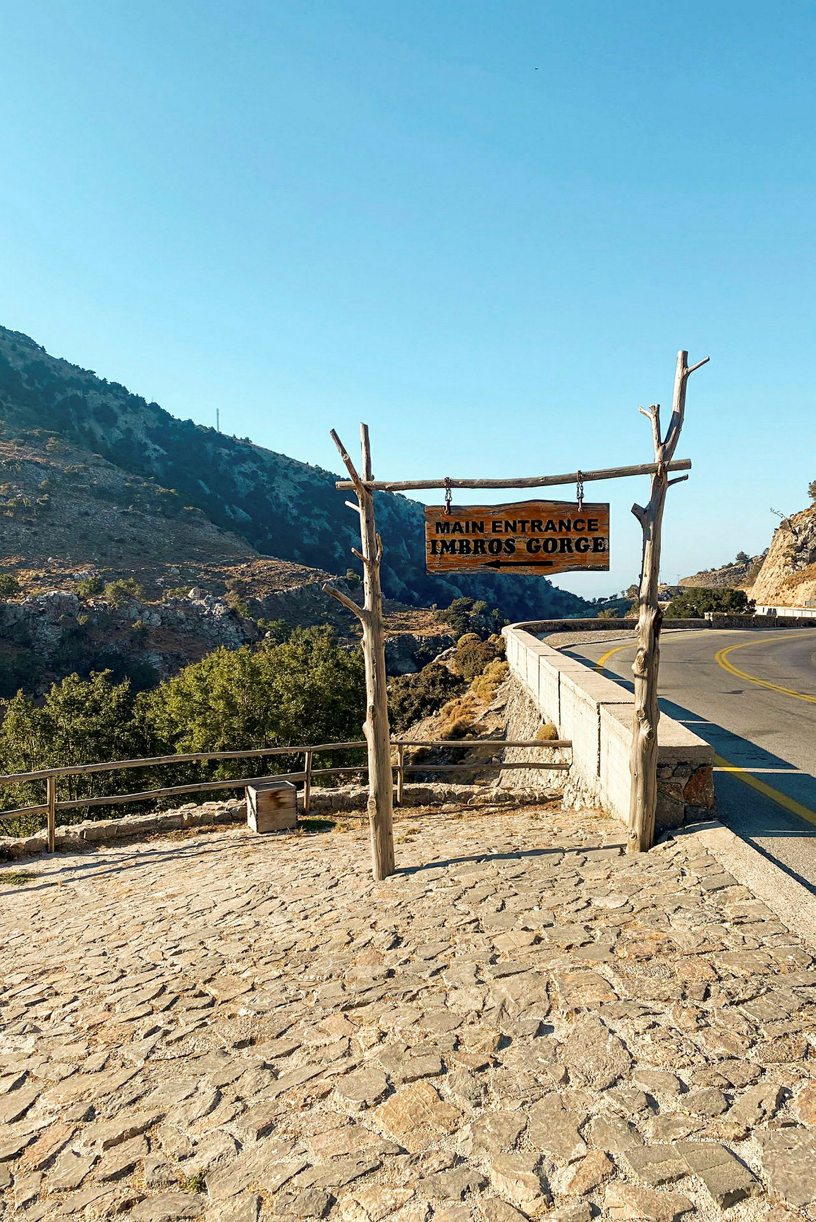 Hovedinngangen til ravinen Imbros Gorge, med et treskilt hengt opp på trestolper, plassert ved siden av en vei og omgitt av fjellandskap. (AI-generert tekst)