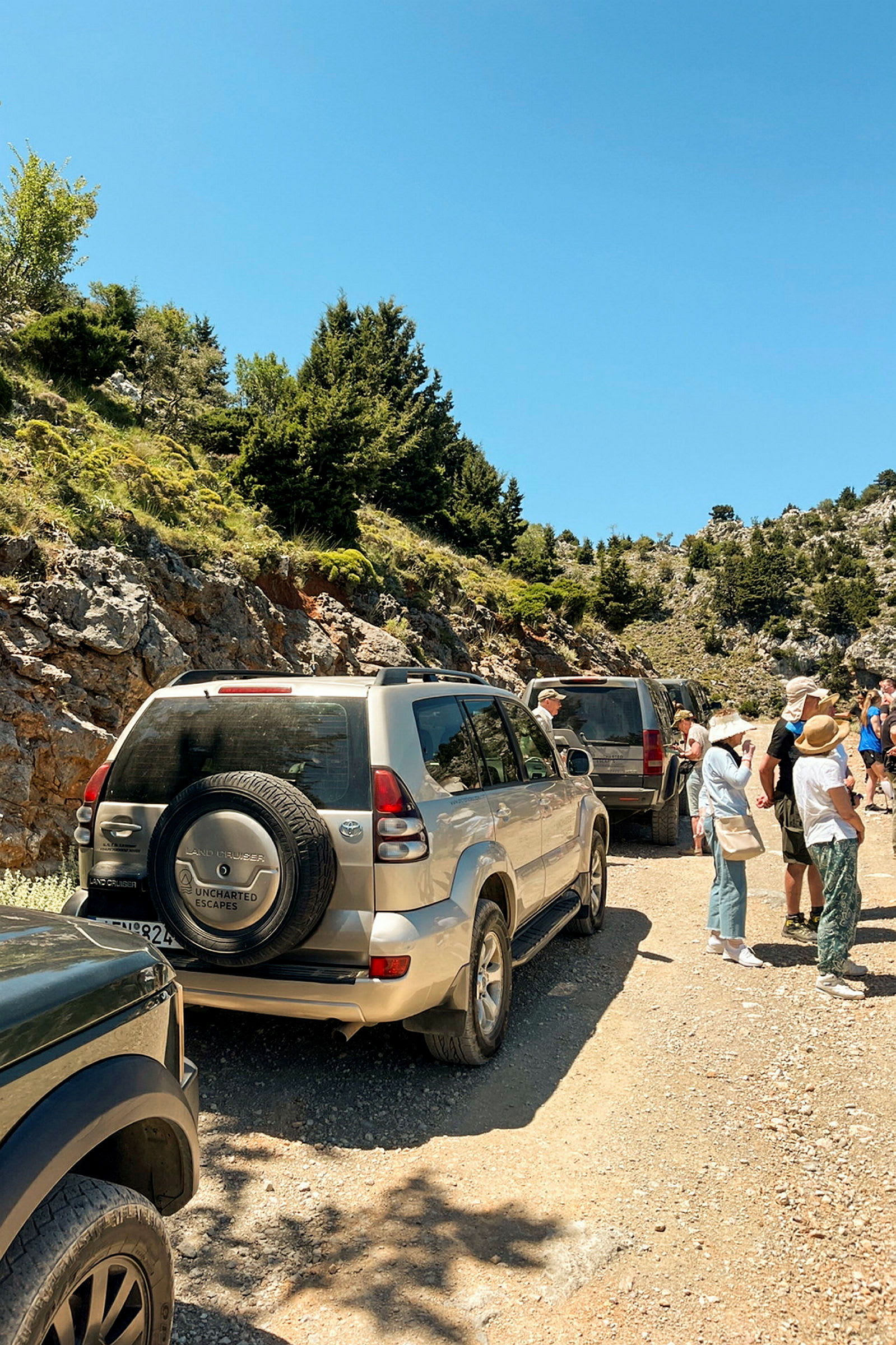 En gruppe terrengbiler står parkert ved en grusvei omgitt av fjellandskap og grønne omgivelser, mens folk samler seg på siden av veien. (AI-generert tekst)
