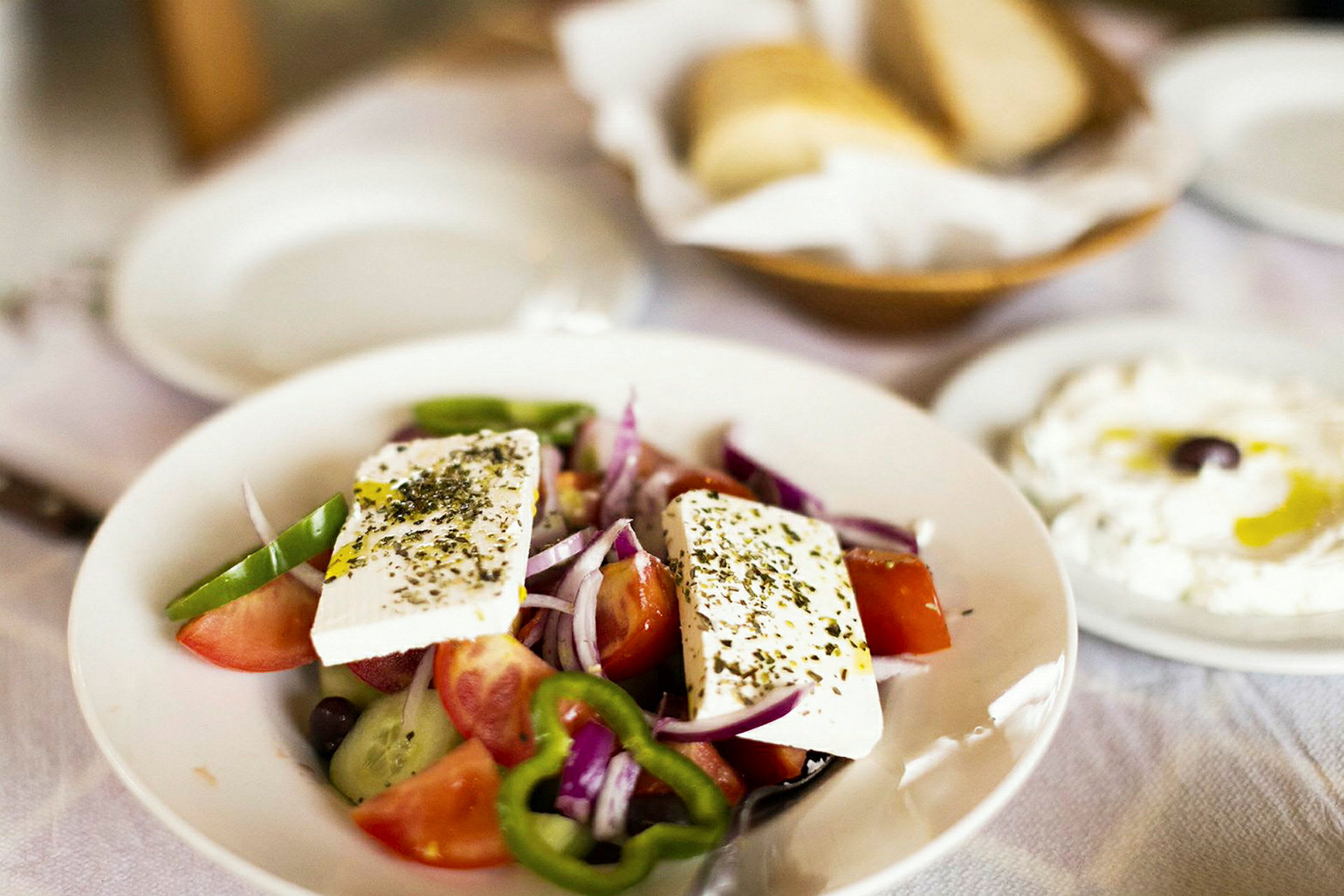 En tallerken med gresk salat som inneholder tomater, agurk, paprika, løk og fetaost. I bakgrunnen sees brød og en tallerken med yoghurt og olivenolje. (AI-generert tekst)