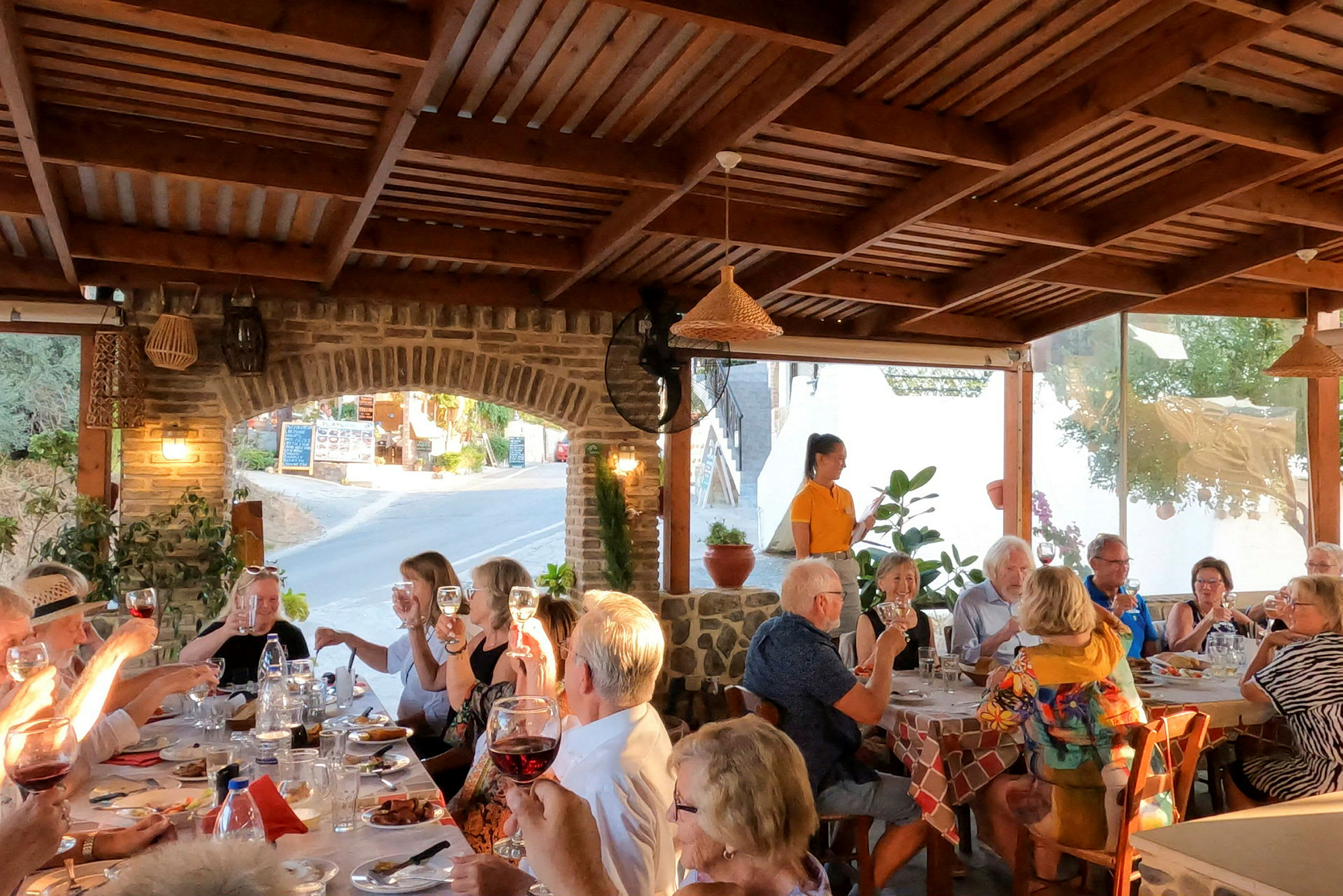 En gruppe mennesker sitter rundt et langbord på en utendørs restaurant under en trepergola. (AI-generert tekst)