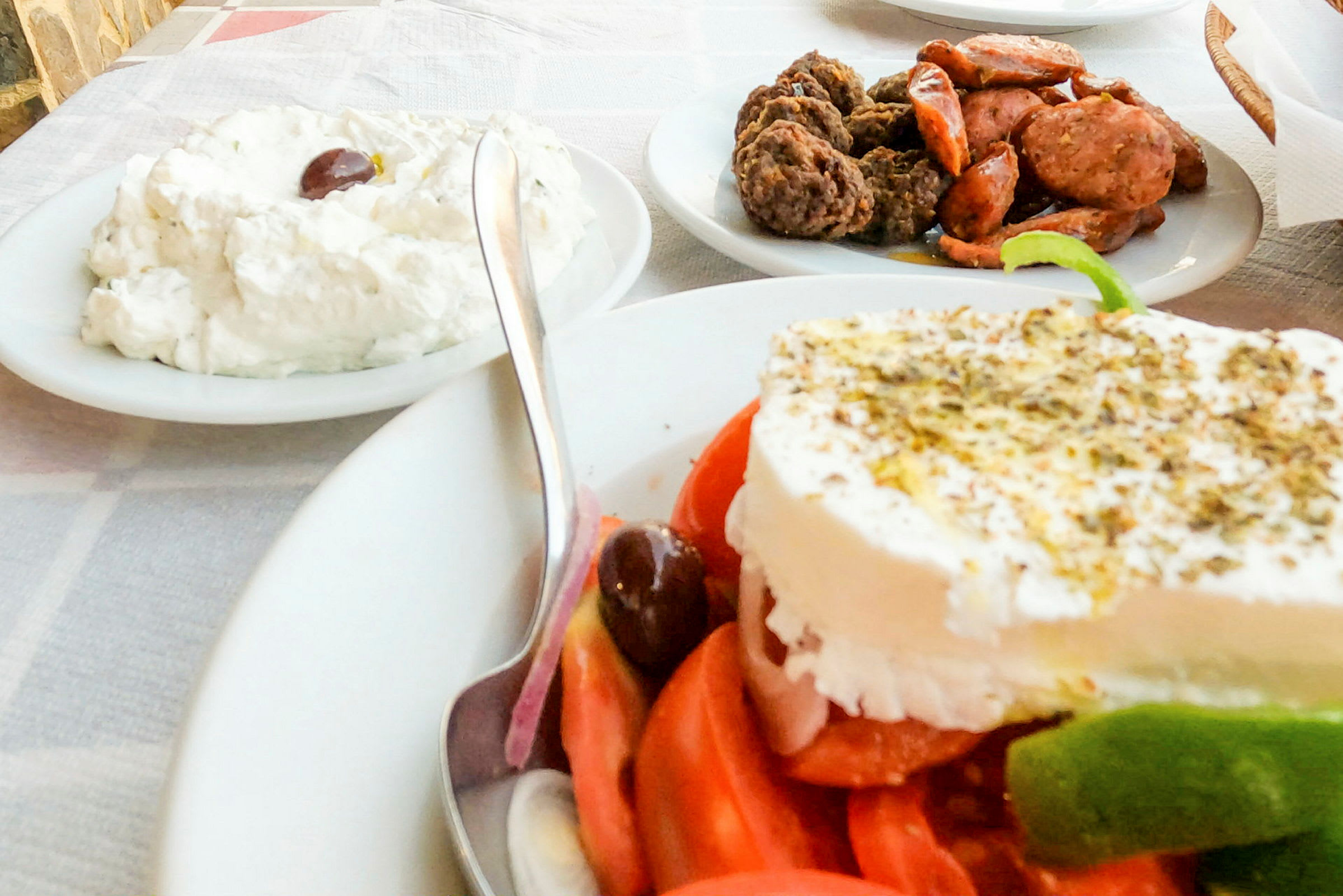 En tallerken med en gresk salat toppet med fetaost, en skål med tzatziki og en tallerken med kjøtt og pølse på et bord. (AI-generert tekst)