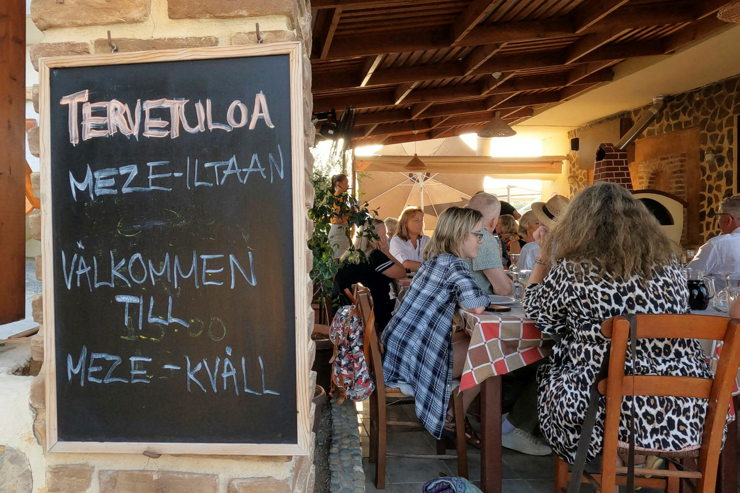 En svart tavle med teksten "Velkommen til meze-kveld" står ved inngangen til en restaurant med gjester sittende ved bord. (AI-generert tekst)