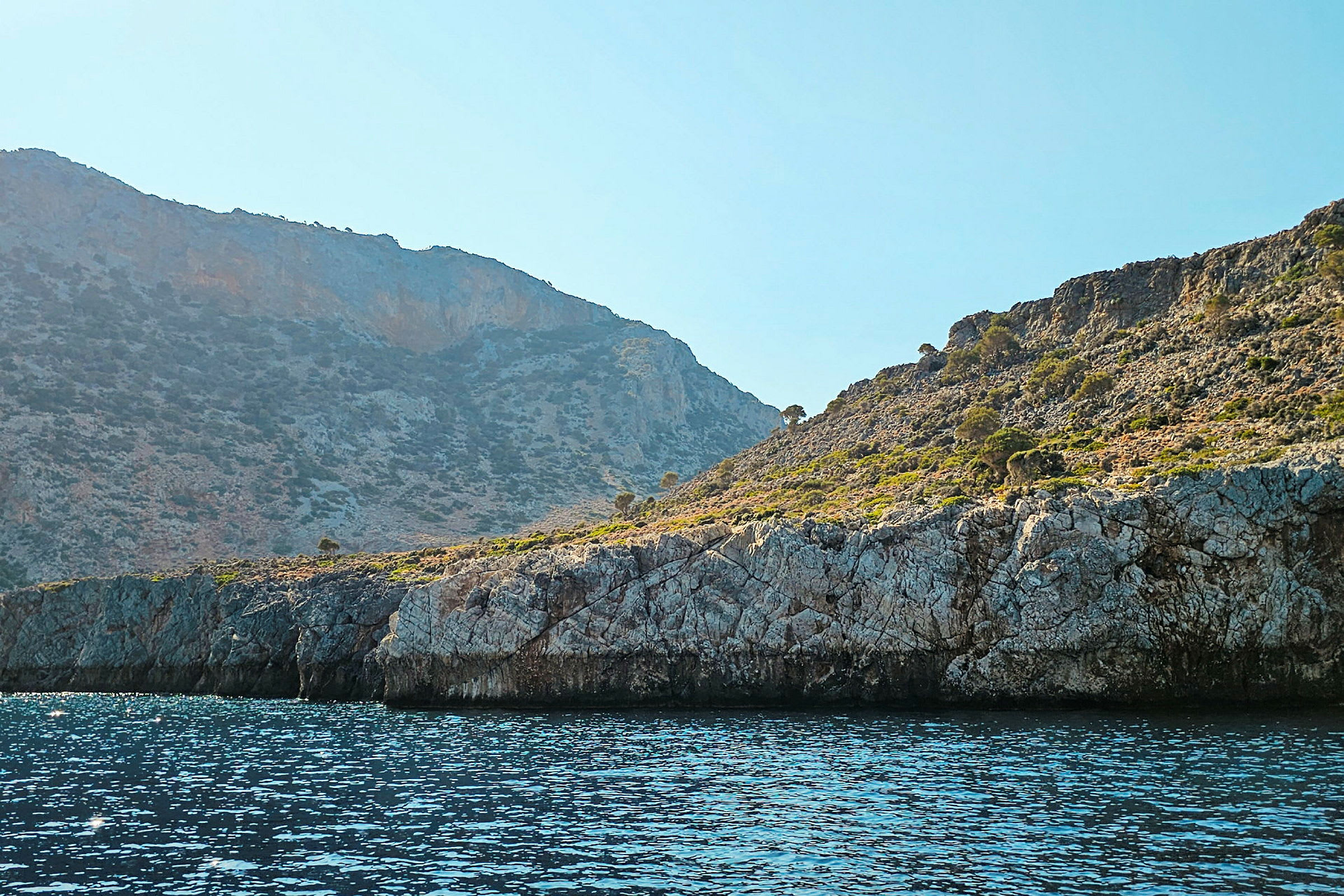 En klippefylt kystlinje med havet i forgrunnen og fjell med vegetasjon i bakgrunnen. (AI-generert tekst)