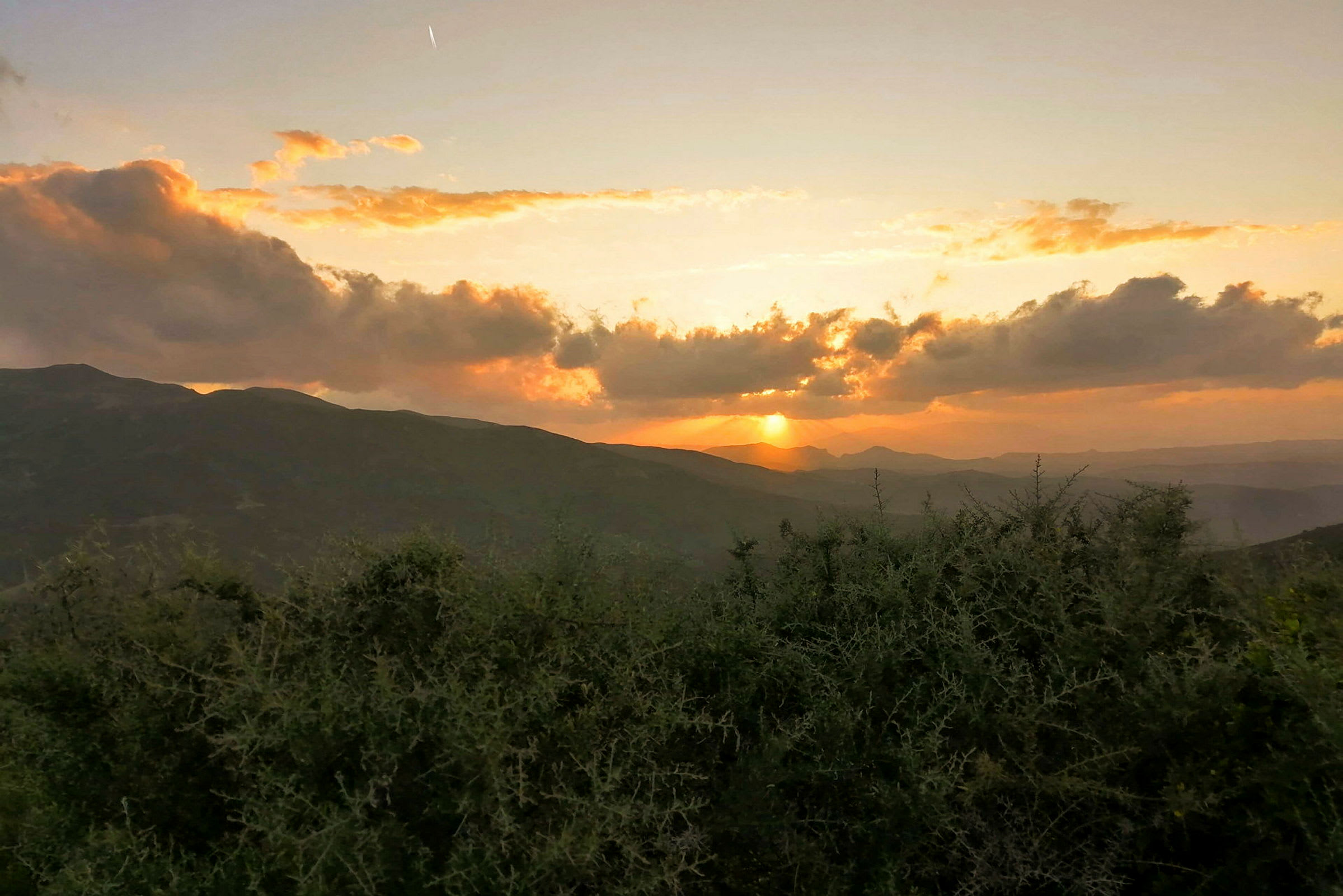 En utsikt over fjell og busker i forgrunnen, med solen som går ned bak horisonten og himmelen farget i gult og oransje toner. (AI-generert tekst)