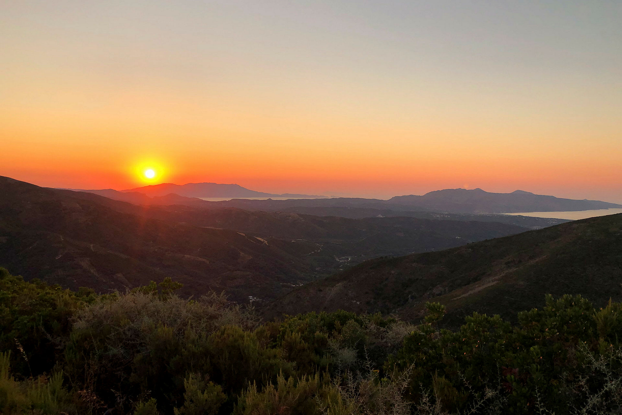 En solnedgang over et fjellandskap med lav vegetasjon og havet i bakgrunnen. (AI-generert tekst)