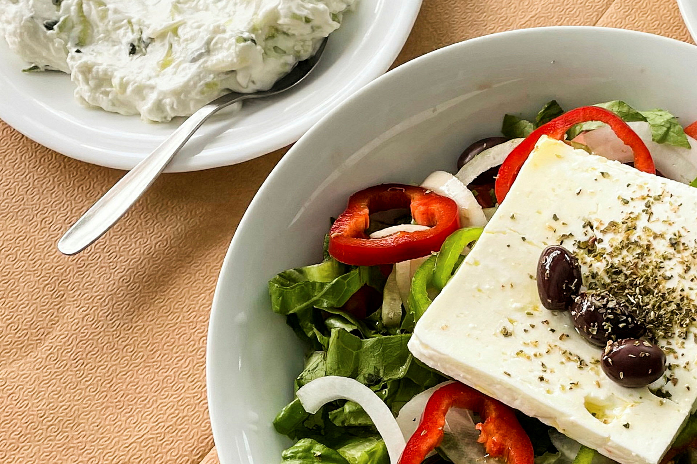 En tallerken med en salat bestående av fetaost, oliven, paprikaringer, løk og salatblader. Ved siden står en skål med tzatziki og en skje. (AI-generert tekst)