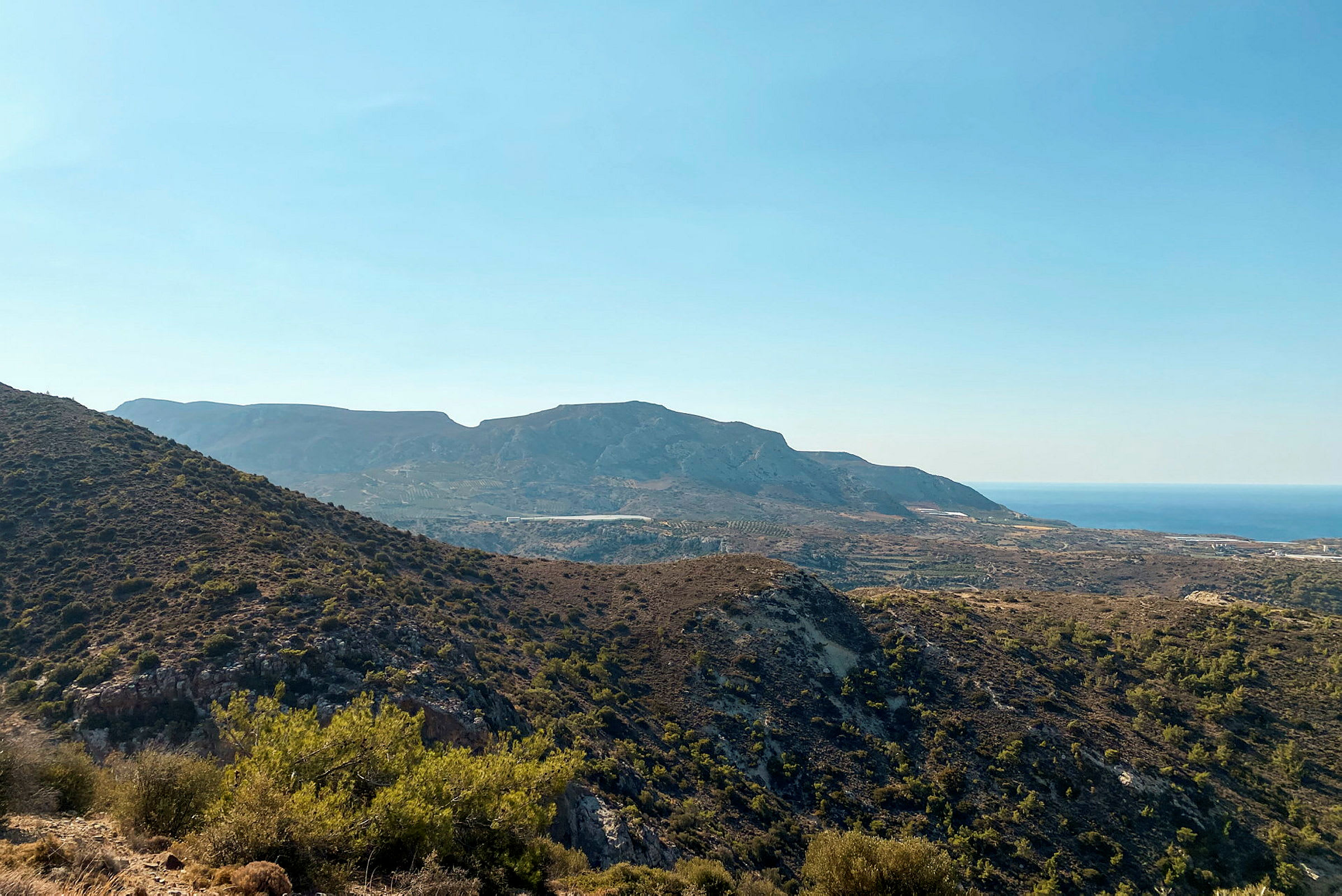 En utsikt over et fjelllandskap med variert vegetasjon og havet i bakgrunnen under en klar blå himmel. (AI-generert tekst)