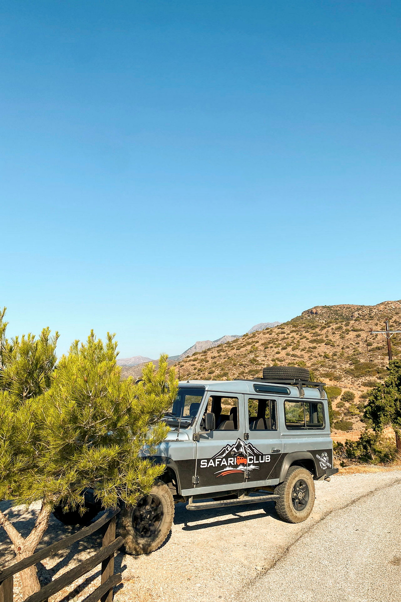 En firehjulsdrevet jeep med teksten "Safari Club" på siden står parkert på en grusvei ved et tre, med fjell og klar himmel i bakgrunnen. (AI-generert tekst)