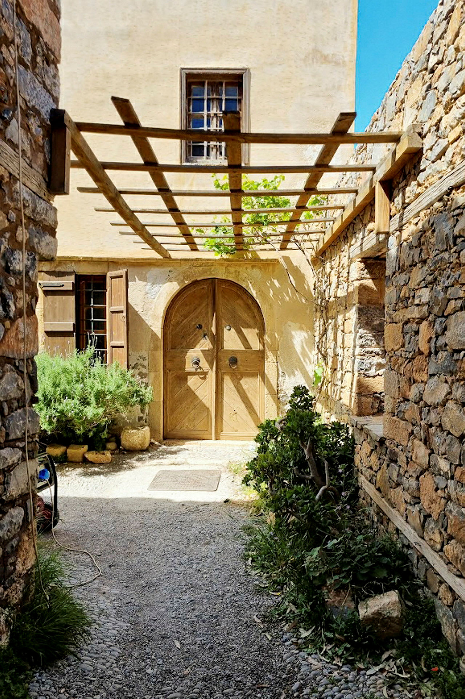 En gammel bygning med en tredør og steinmur omgitt av planter under en pergola. (AI-generert tekst)