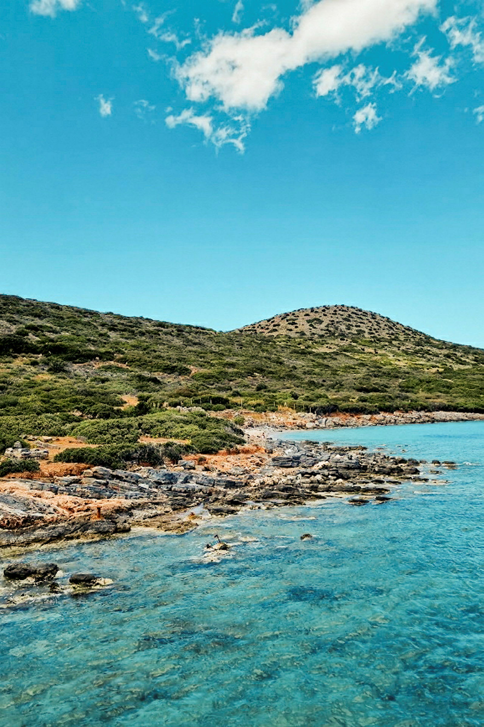 Kystlinje med klipper og buskvegetasjon, samt klart turkist havvann under en blå himmel. (AI-generert tekst)