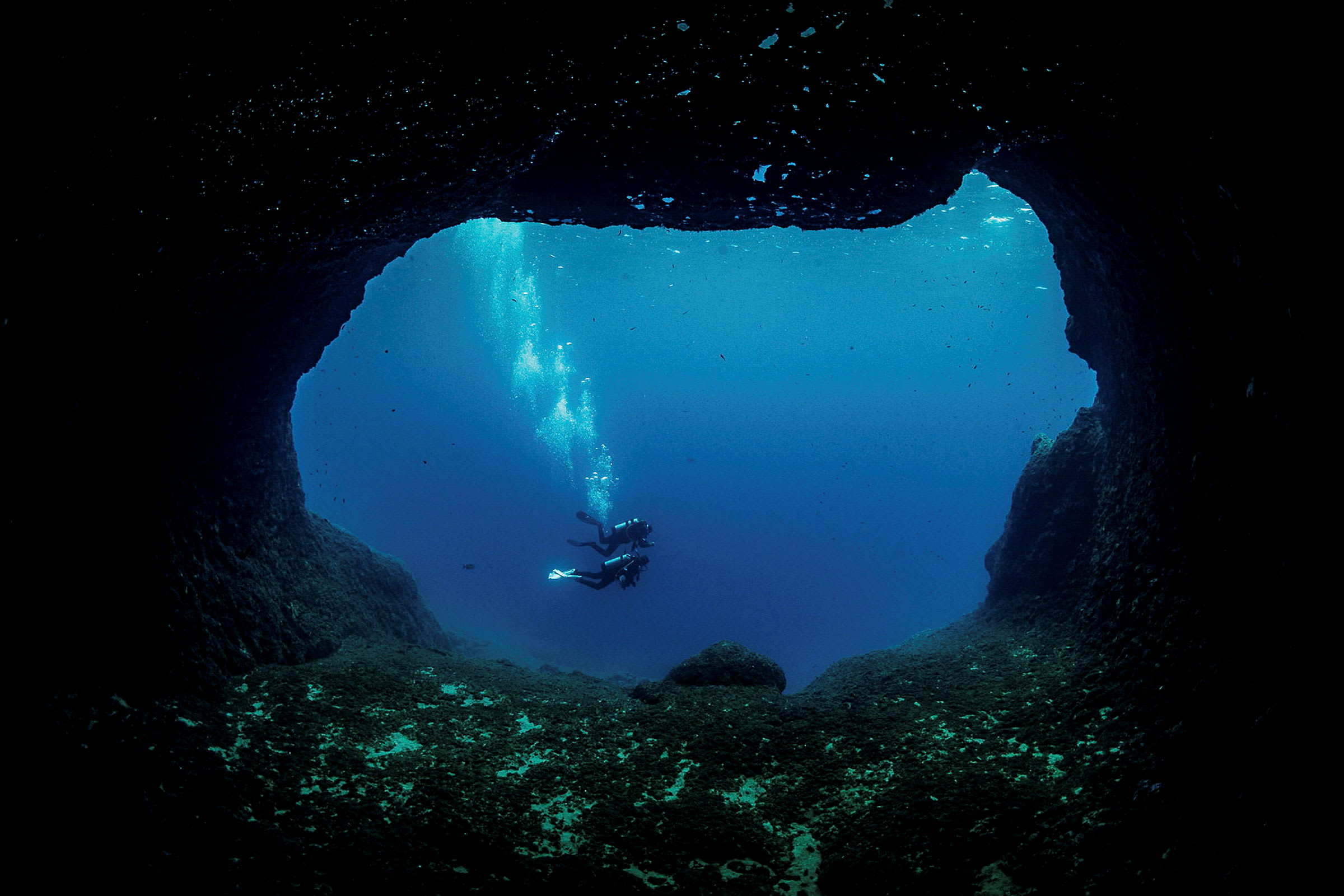 To dykkere svømmer i havet, sett fra innsiden av en undervannsgrotte. (AI-generert tekst)