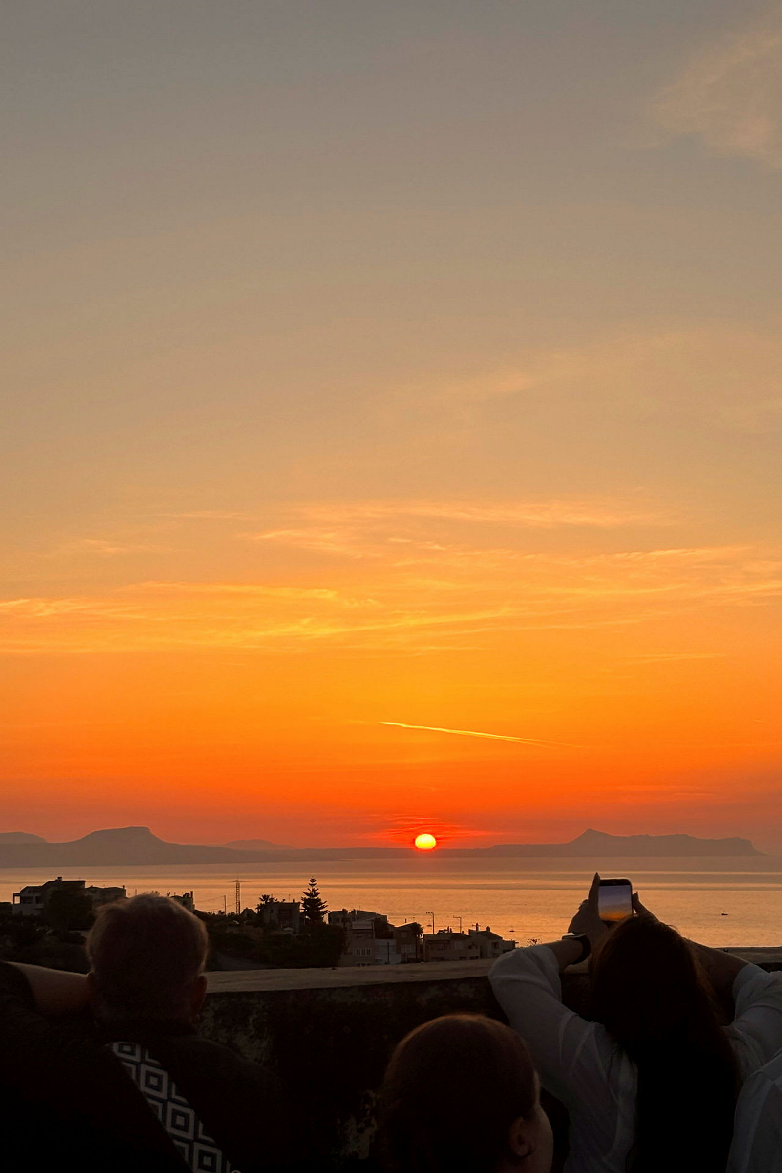 En solnedgang over havet, med silhuetter av bygninger og personer som observerer og fotograferer utsikten. (AI-generert tekst)
