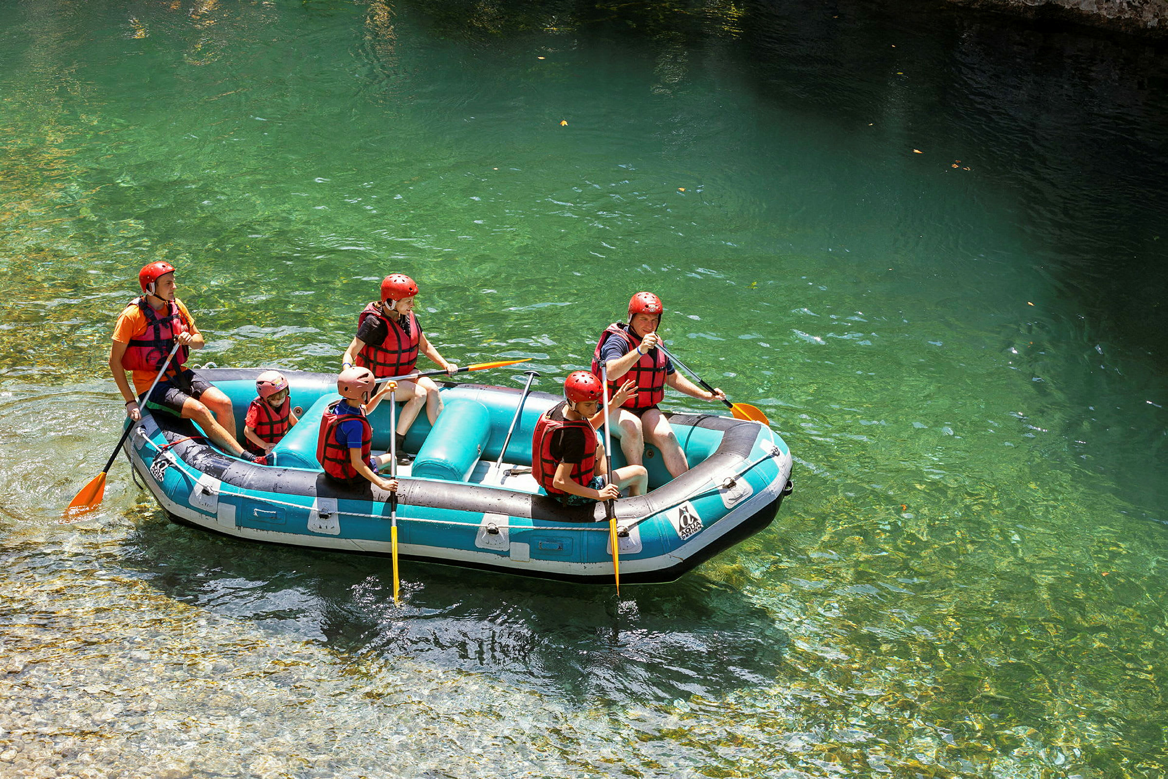 En gruppe mennesker driver med rafting i en oppblåsbar flåte på klart vann, og har på seg hjelmer og redningsvester. (AI-generert tekst)
