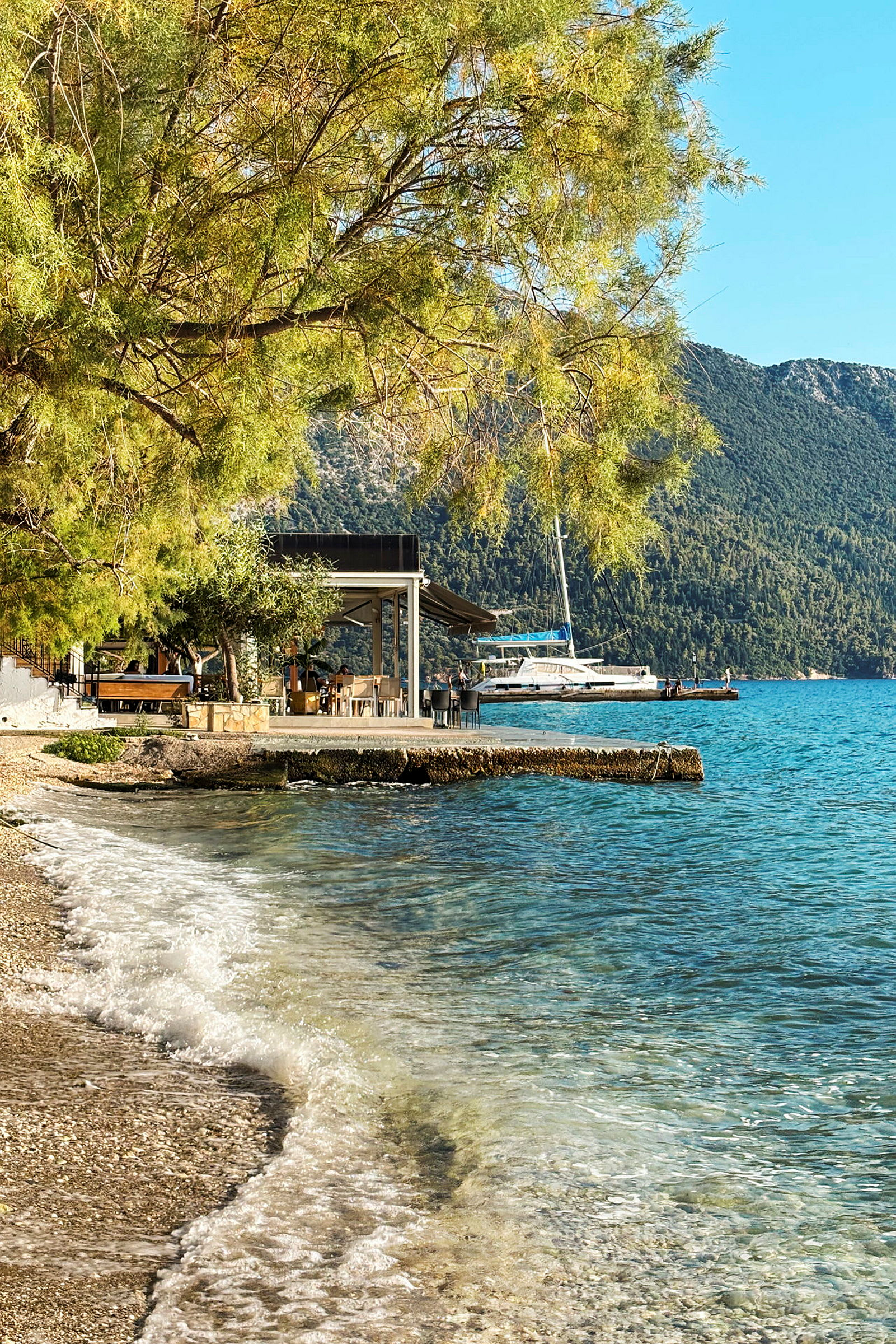 Strand med klart vann og bølger som skyller mot kysten. En terrasse med bord og stoler ses ved vannkanten, samt en seilbåt i bakgrunnen. (AI-generert tekst)