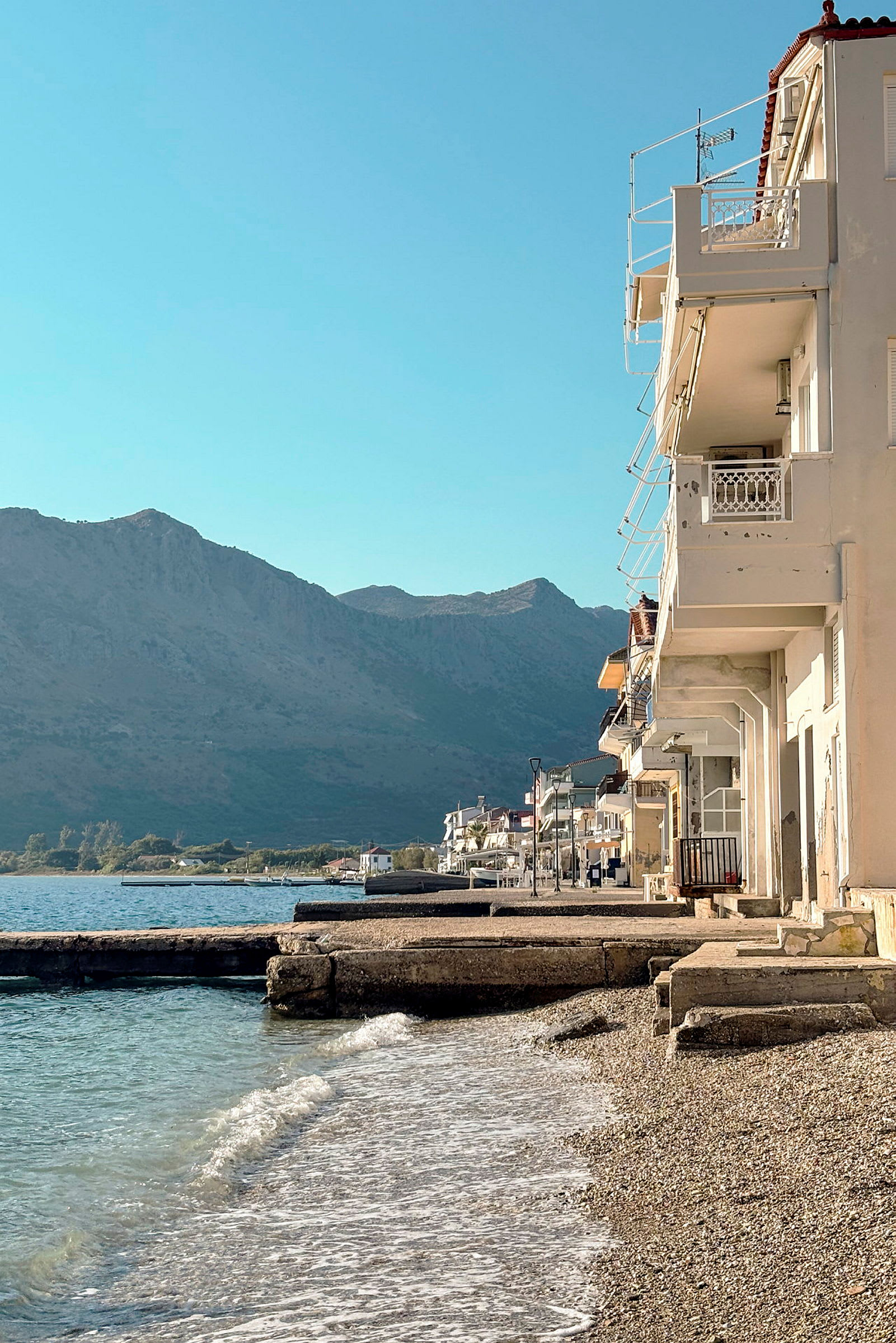 Utsikt over strandpromenade med bygninger ved stranden og fjell i bakgrunnen. (AI-generert tekst)