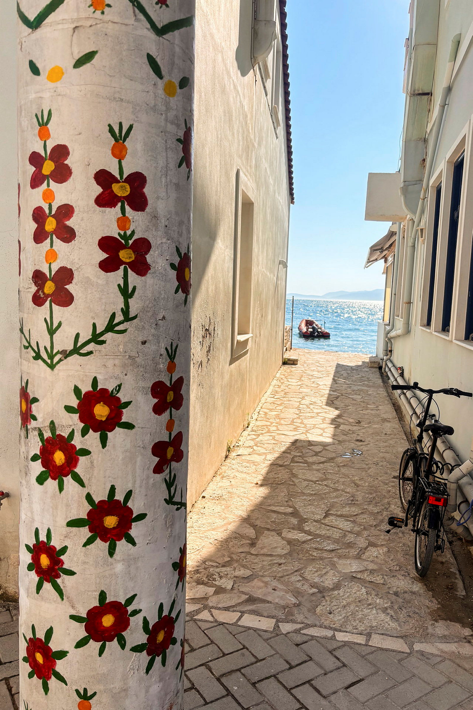 En smal steinlagt passasje mellom hus, med en søyle dekorert med malte røde blomster i forgrunnen og havet synlig i bakgrunnen. (AI-generert tekst)