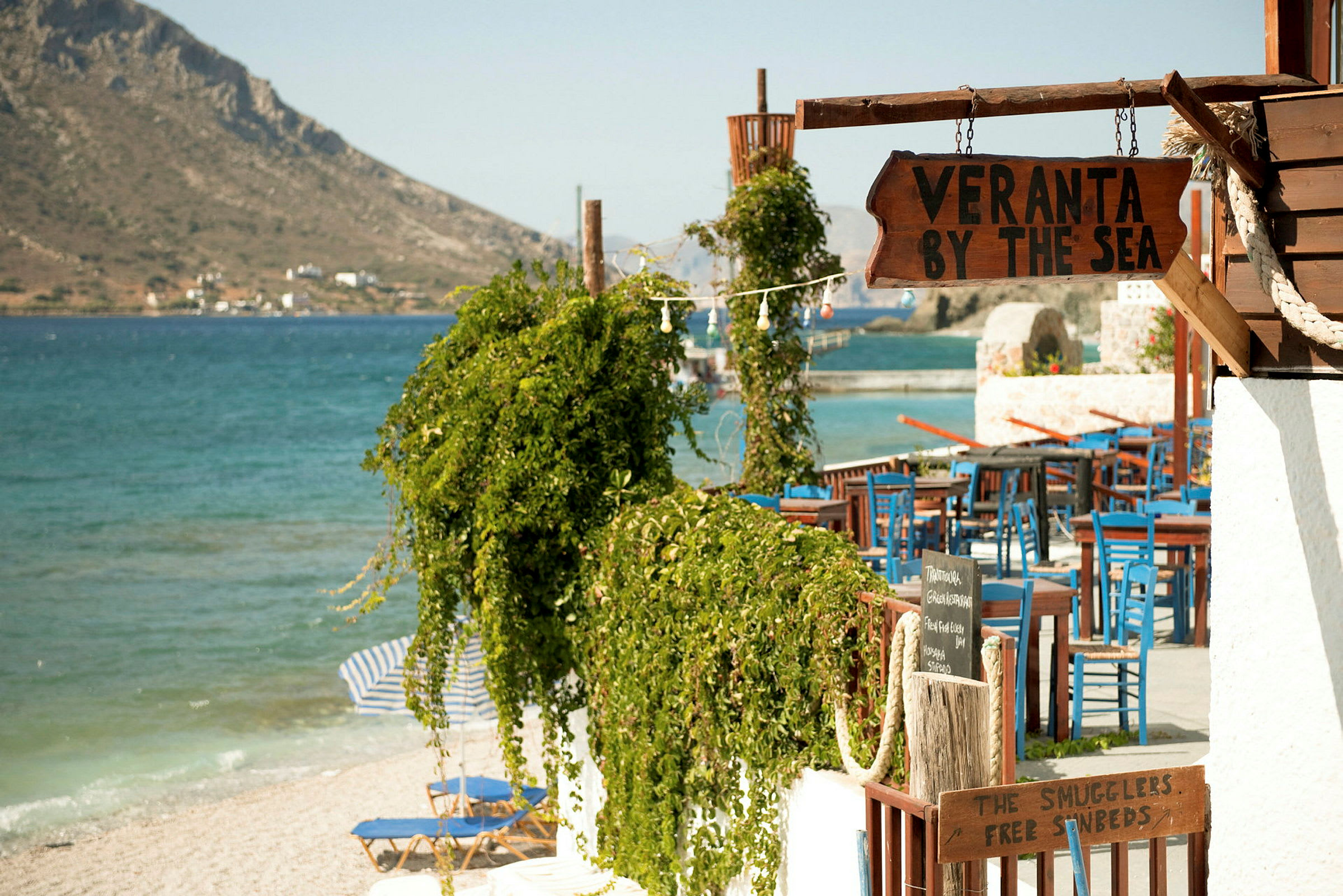 Et nærbilde av en restaurant eller veranda ved stranden med skilt som viser "Veranta by the sea" og blå stoler samt et grønt område i forgrunnen. (AI-generert tekst)