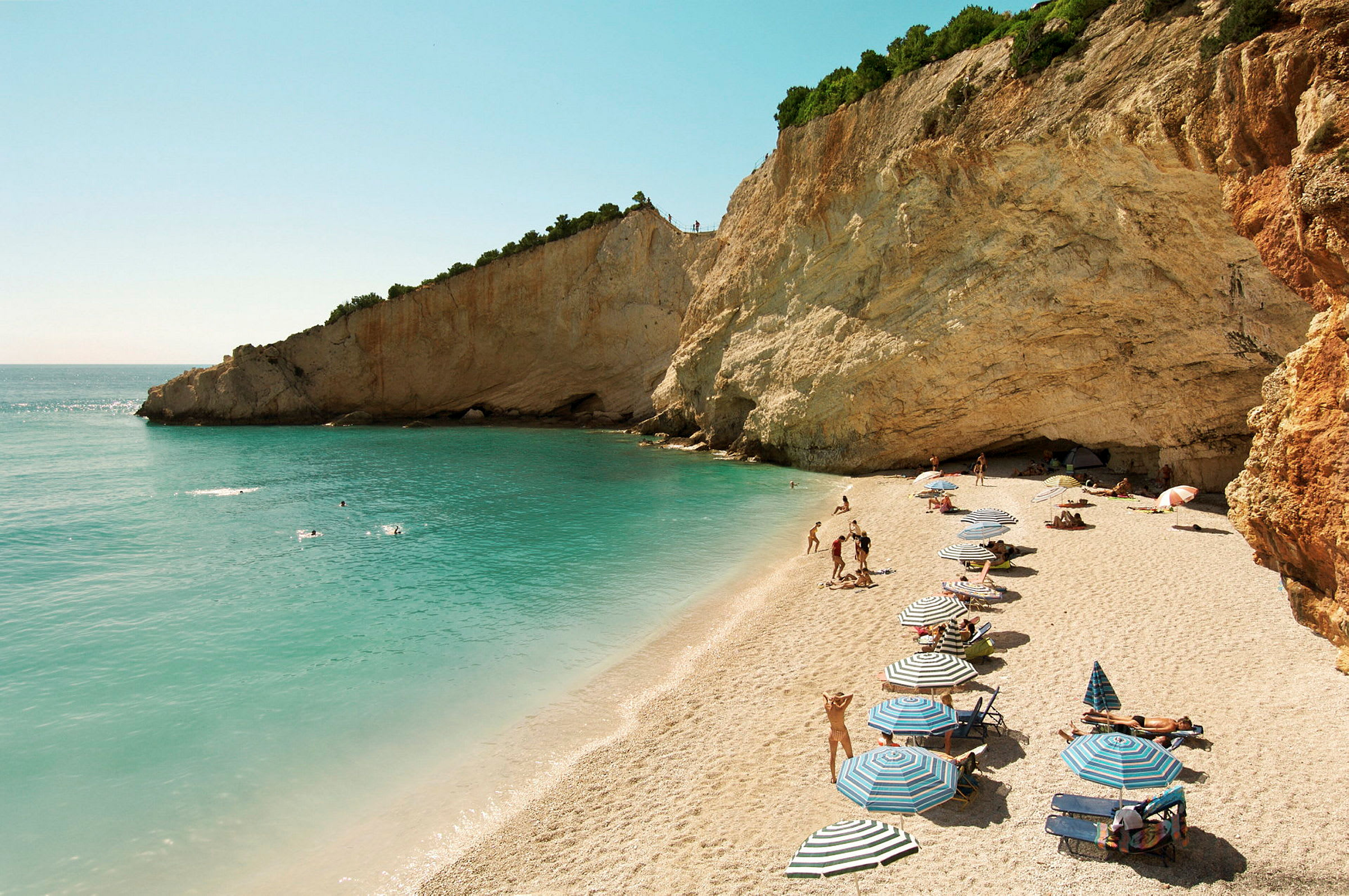 Strand med parasoller, sand, klippevegg og svømmende mennesker ved et turkist hav. (AI-generert tekst)