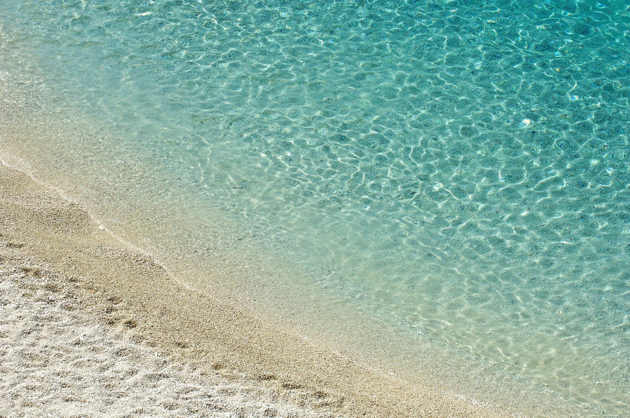 Bildet viser en strandkant der klart blått vann møter lys sand. (AI-generert tekst)