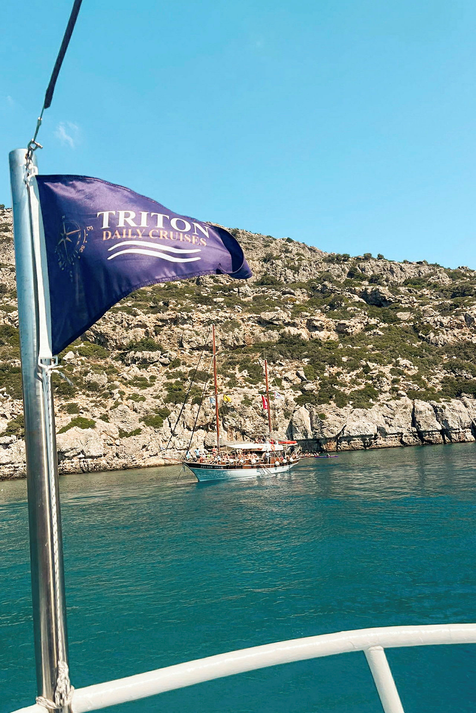 Et seilskip ligger ved en klippe mens et flagg med teksten "Triton Daily Cruises" vaier i forgrunnen. (AI-generert tekst)
