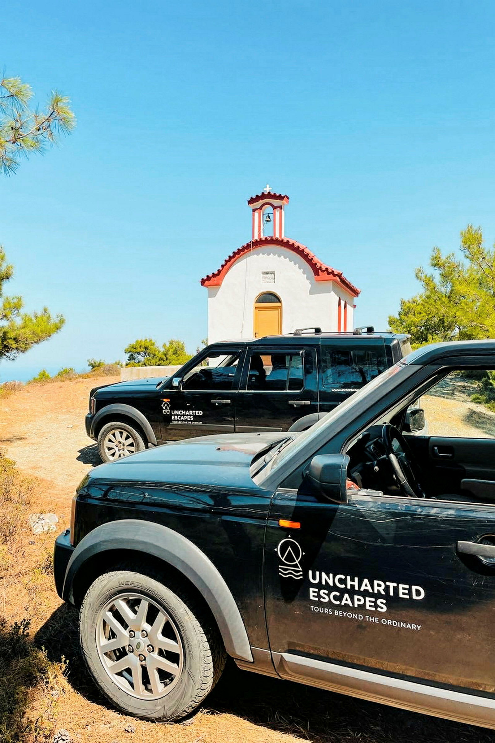 To svarte biler med logoen "Uncharted Escapes" står parkert foran en liten, hvit kirke med rødt tak. Kirken er omgitt av grøntområder, og himmelen er klar og blå. (AI-generert tekst)