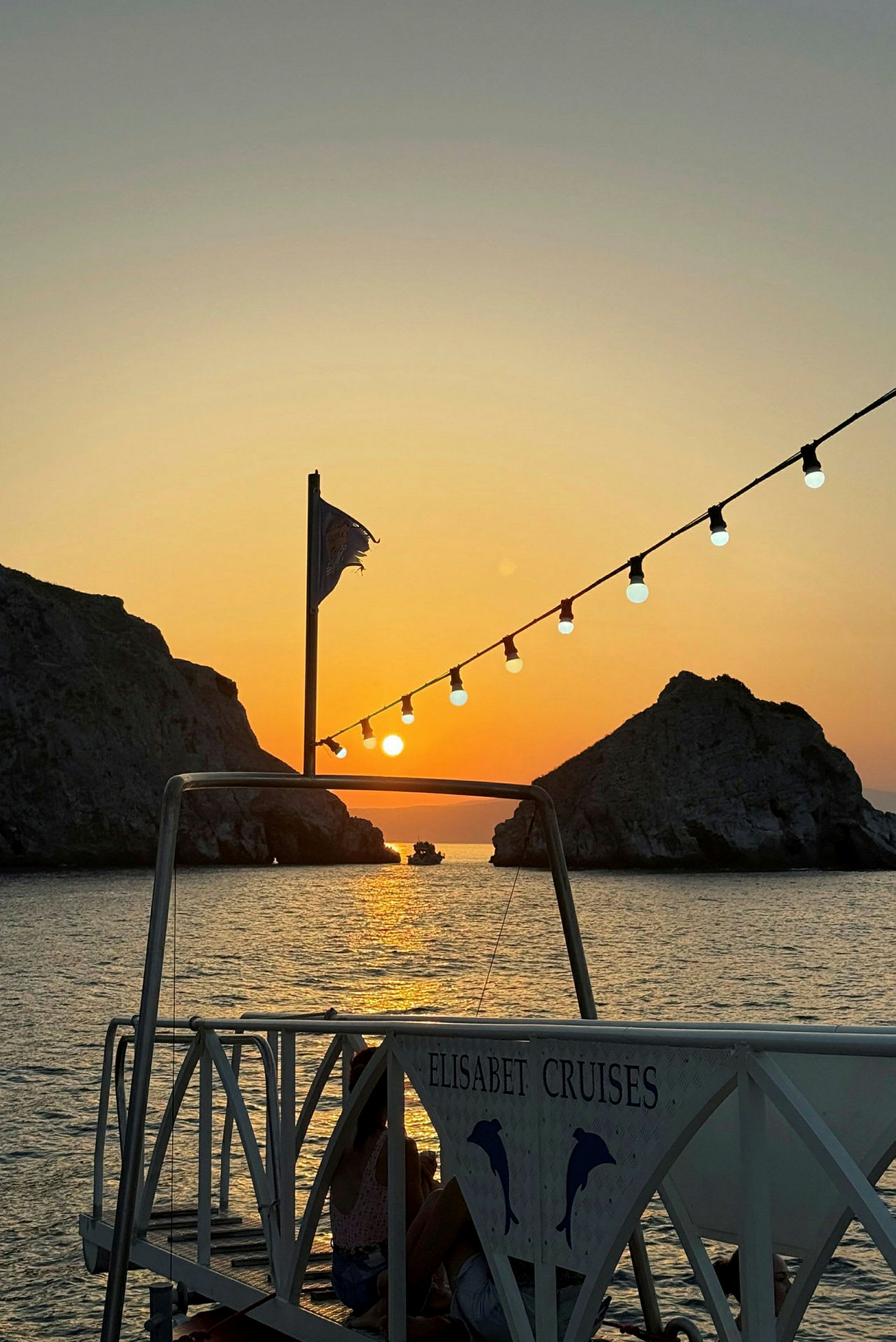 En solnedgang over havet med en brygge i forgrunnen som har et skilt med teksten 'Elisabet Cruises'. I bakgrunnen er det klipper og en båt. (AI-generert tekst)
