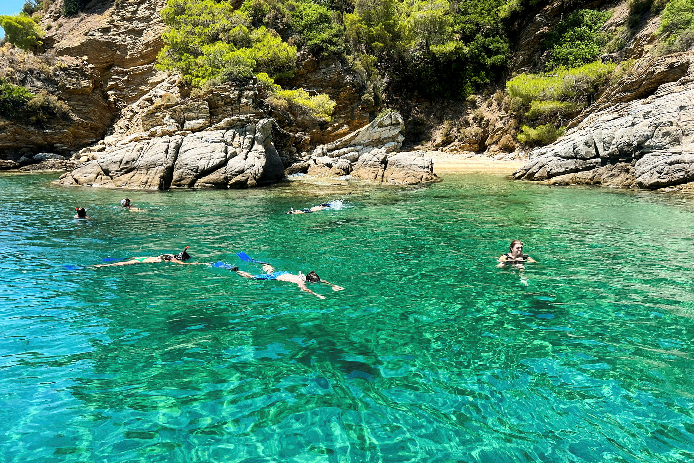 Personer svømmer og snorkler i klart turkis vann nær en steinete strand. (AI-generert tekst)