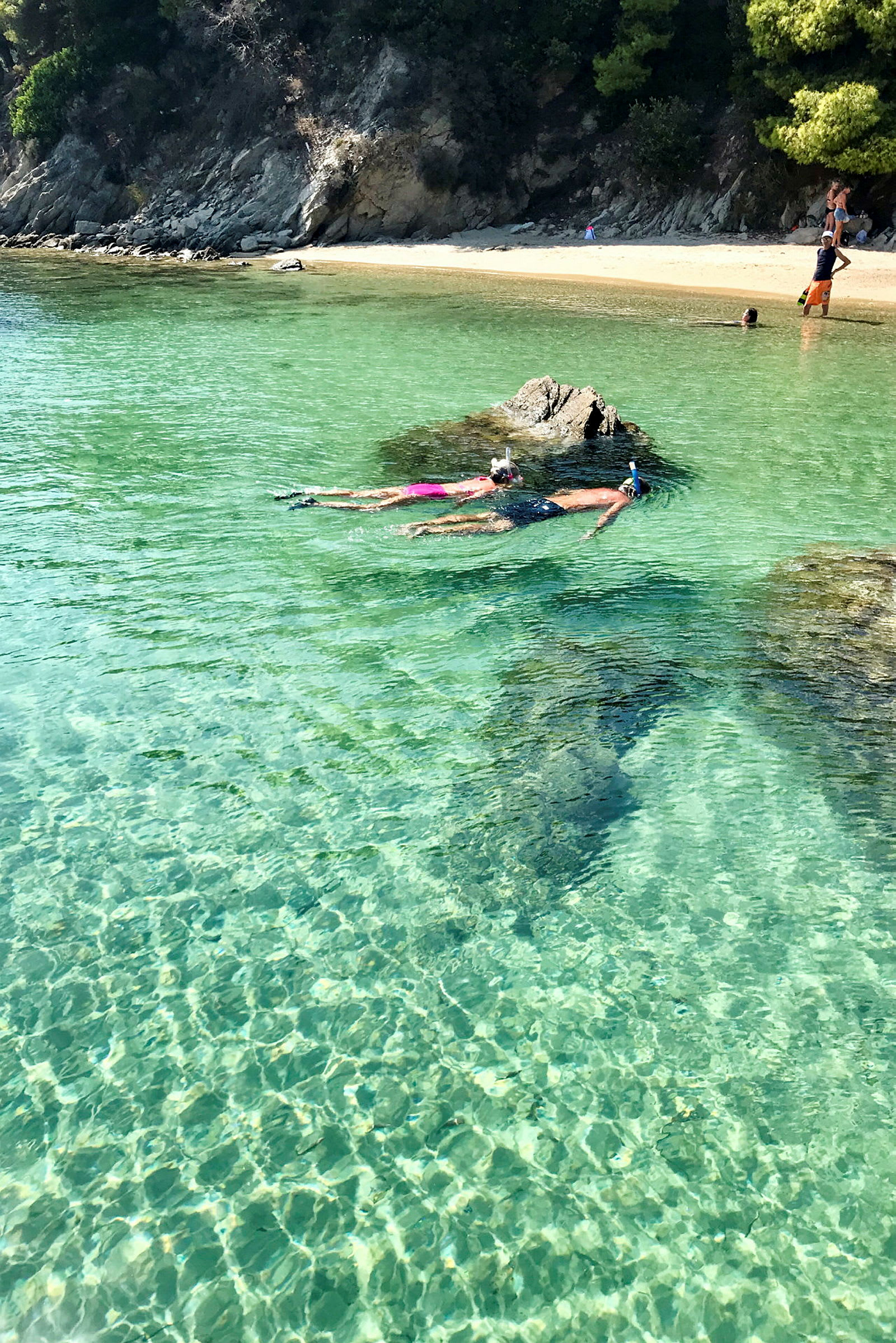 To personer snorkler i klart, turkis vann nær en stein, med en sandstrand og trær i bakgrunnen. (AI-generert tekst)