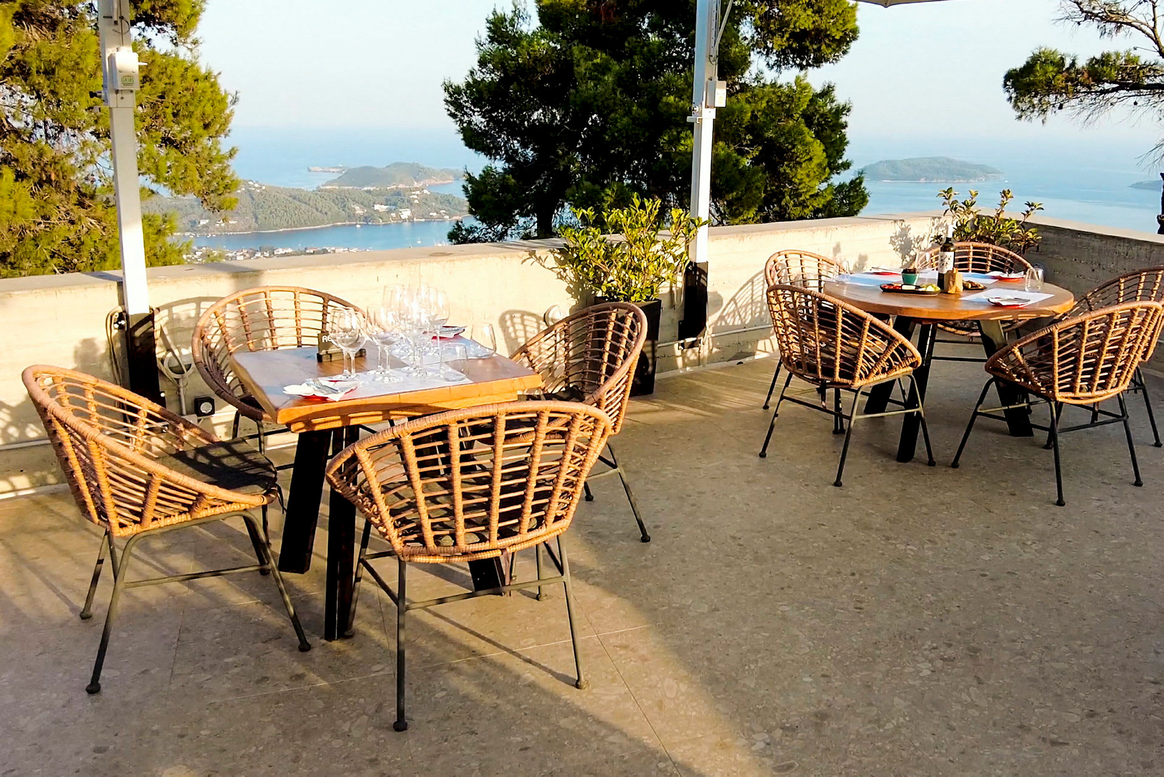 Utendørs restaurant med bord og flettede stoler, med utsikt over havet og grønne omgivelser. (AI-generert tekst)