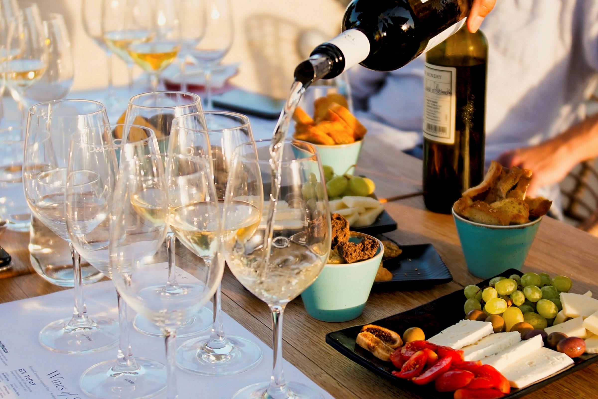 Et bord med glass fylt med hvitvin som serveres, omgitt av ulike snacks som ost, brød og frukt. (AI-generert tekst)