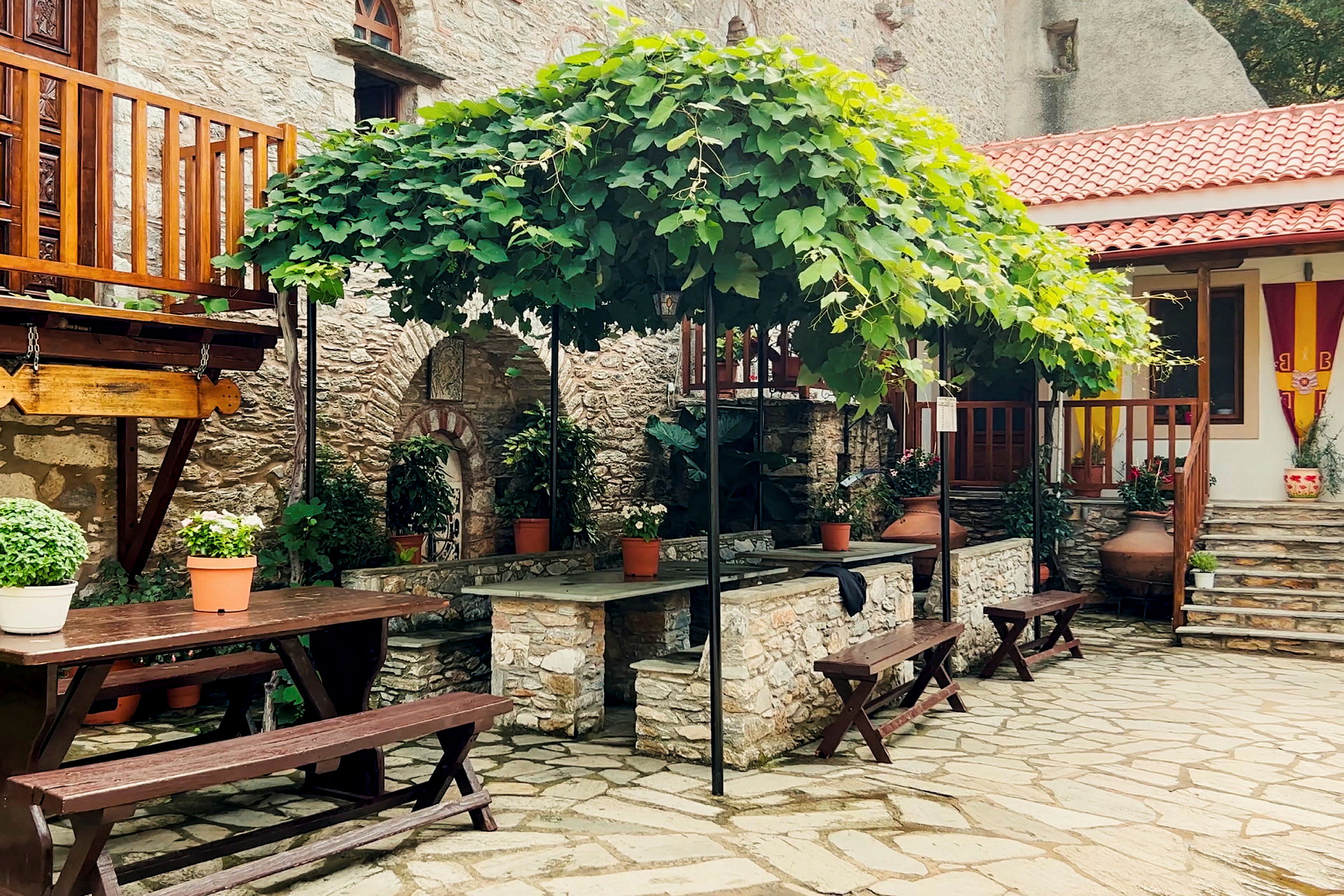 En uteplass med steinlagt gulv, trebord og benker samt store blomsterkrukker. Grønne planter klatrer på en pergola over. (AI-generert tekst)