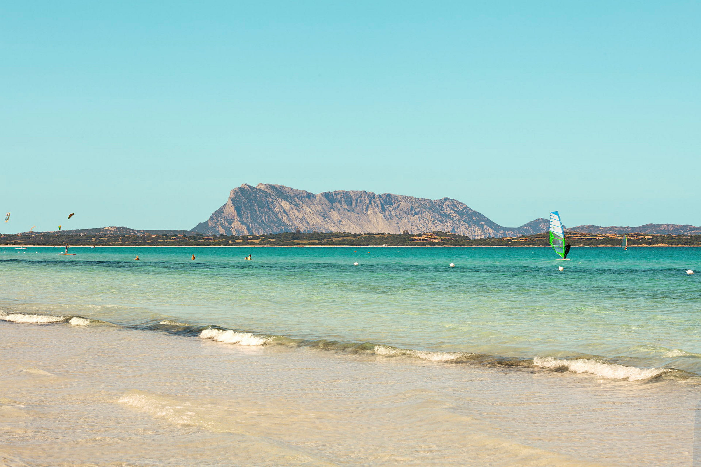 En strand med klart vann, folk bader og en person windsurfer. En stor fjellformasjon kan sees i bakgrunnen. (AI-generert tekst)