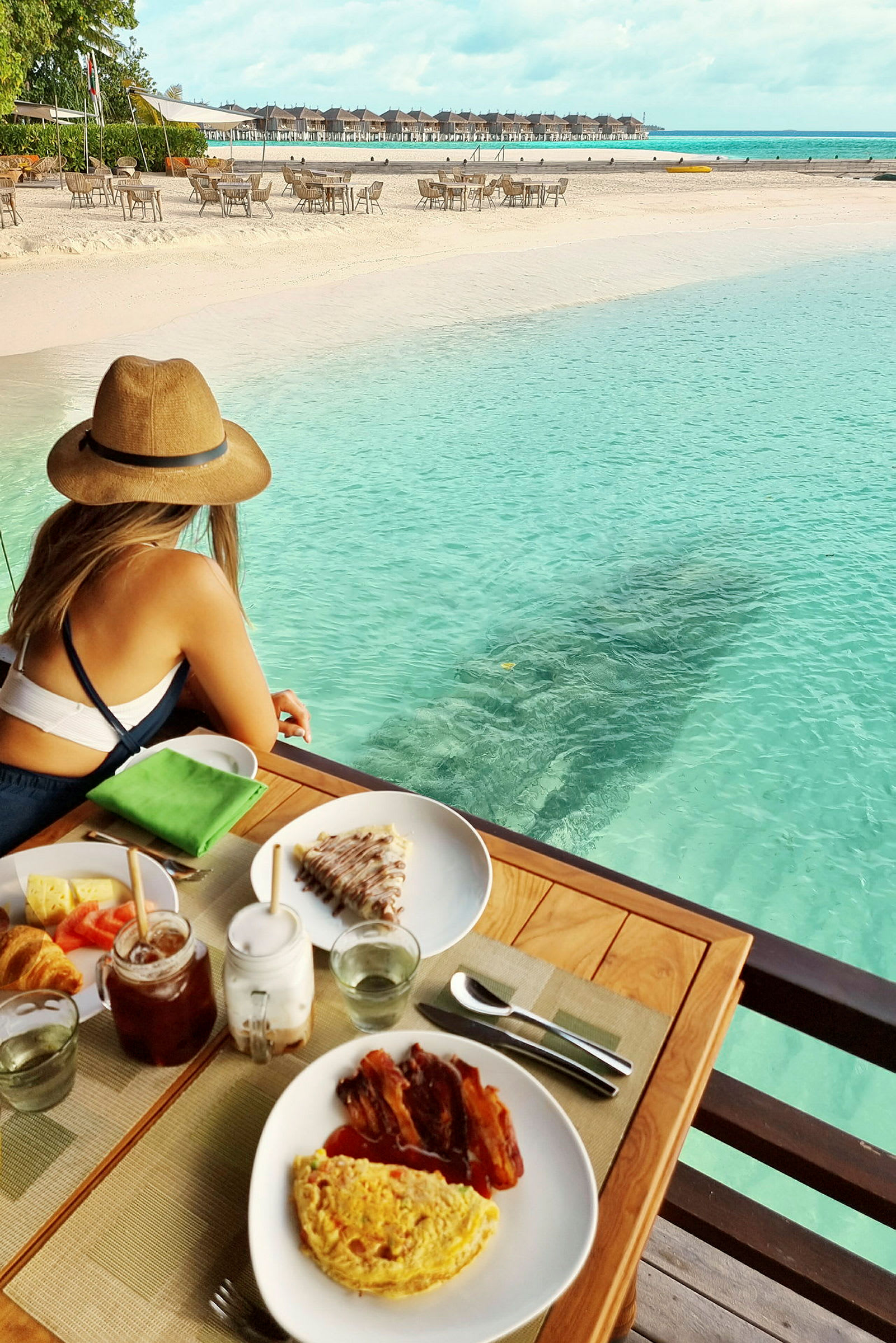 En kvinne sitter ved et bord med frokostmat, ved strandkanten med turkist vann og utsikt over stranden og bungalower. (AI-generert tekst)