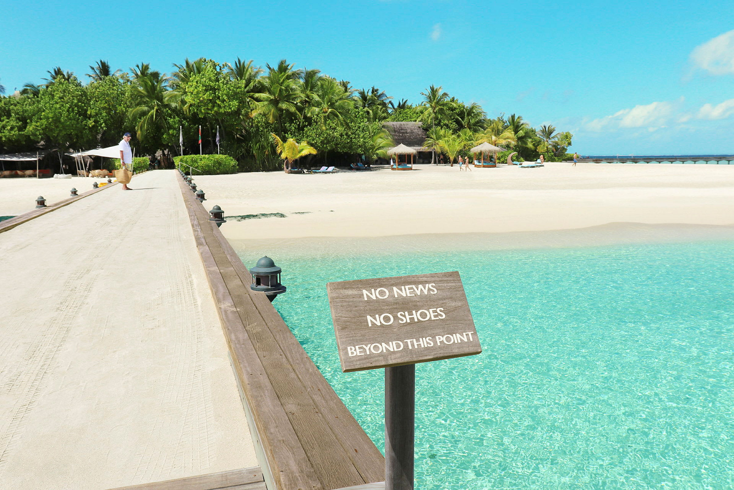 En sandete gangbro fører til en tropisk strand med et skilt som lyder 'No News No Shoes Beyond This Point'. (AI-generert tekst)