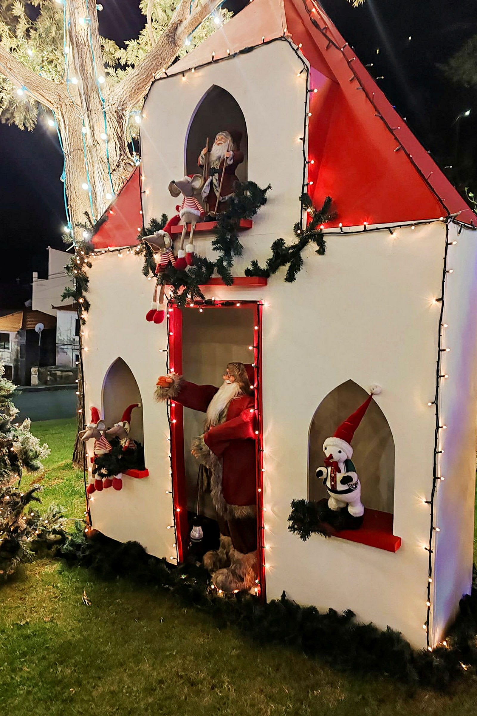 En juledekorasjon som forestiller et hus med julenissen og leketøy, dekorert med lyslenker og kranser. (AI-generert tekst)