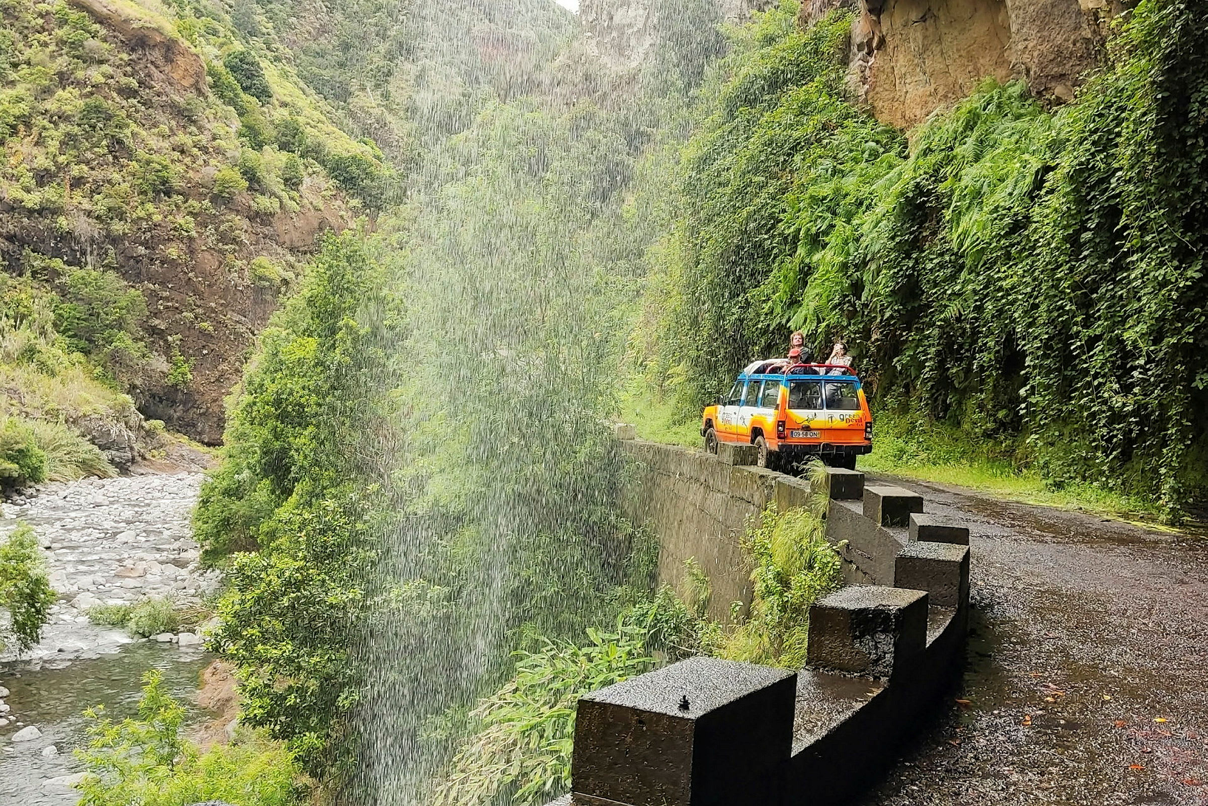 En oransje jeep parkert på en smal vei ved siden av en grønn dal med regn eller vannsprut. (AI-generert tekst)