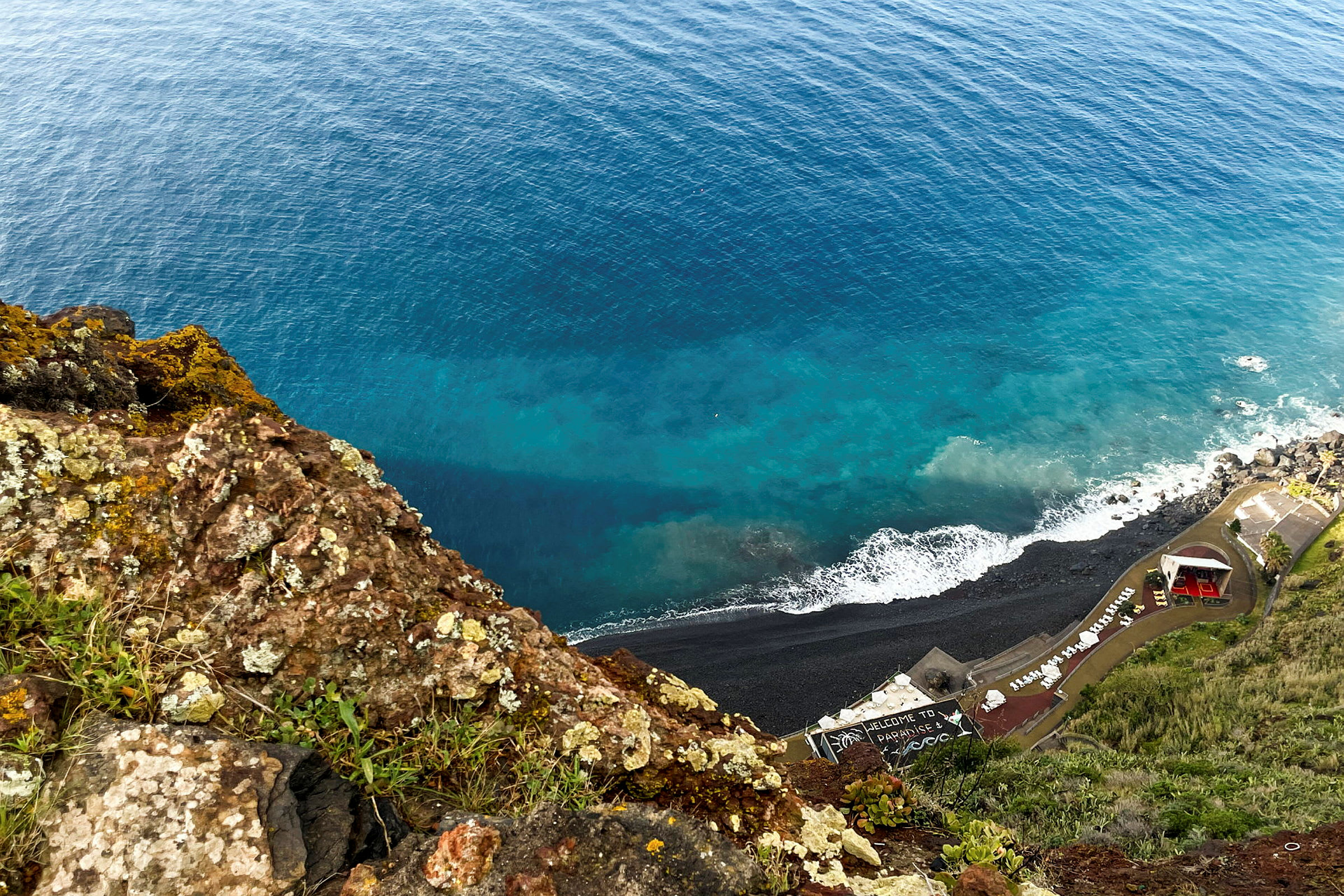Utsikt fra en klippe ned mot en kystlinje med steinstrand og bygninger, samt havet. (AI-generert tekst)