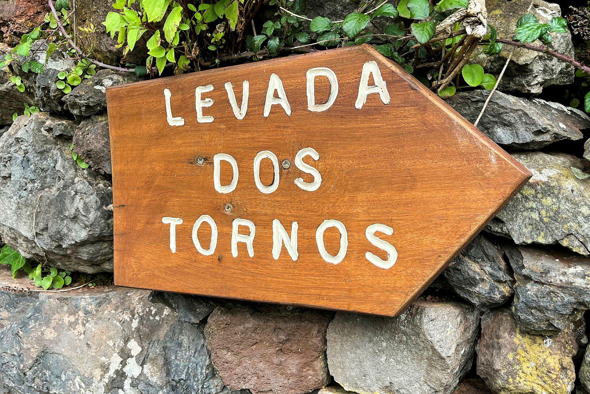 Et treskilt med teksten 'Levada dos Tornos' plassert mot en bakgrunn av steiner og planter. (AI-generert tekst)