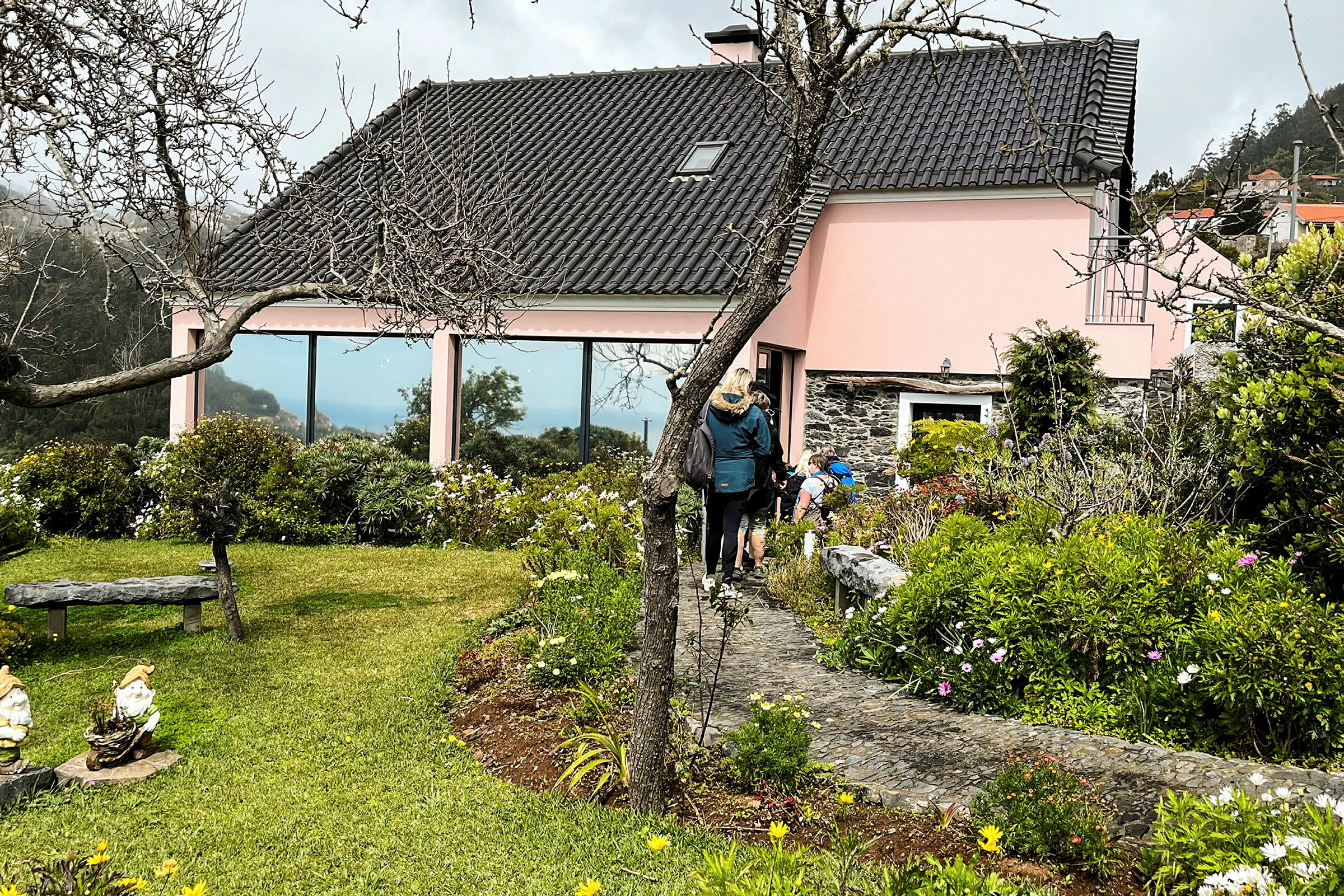 En rosa bygning med svart tak omgitt av grønt og blomster, med personer som går på en gangvei. (AI-generert tekst)