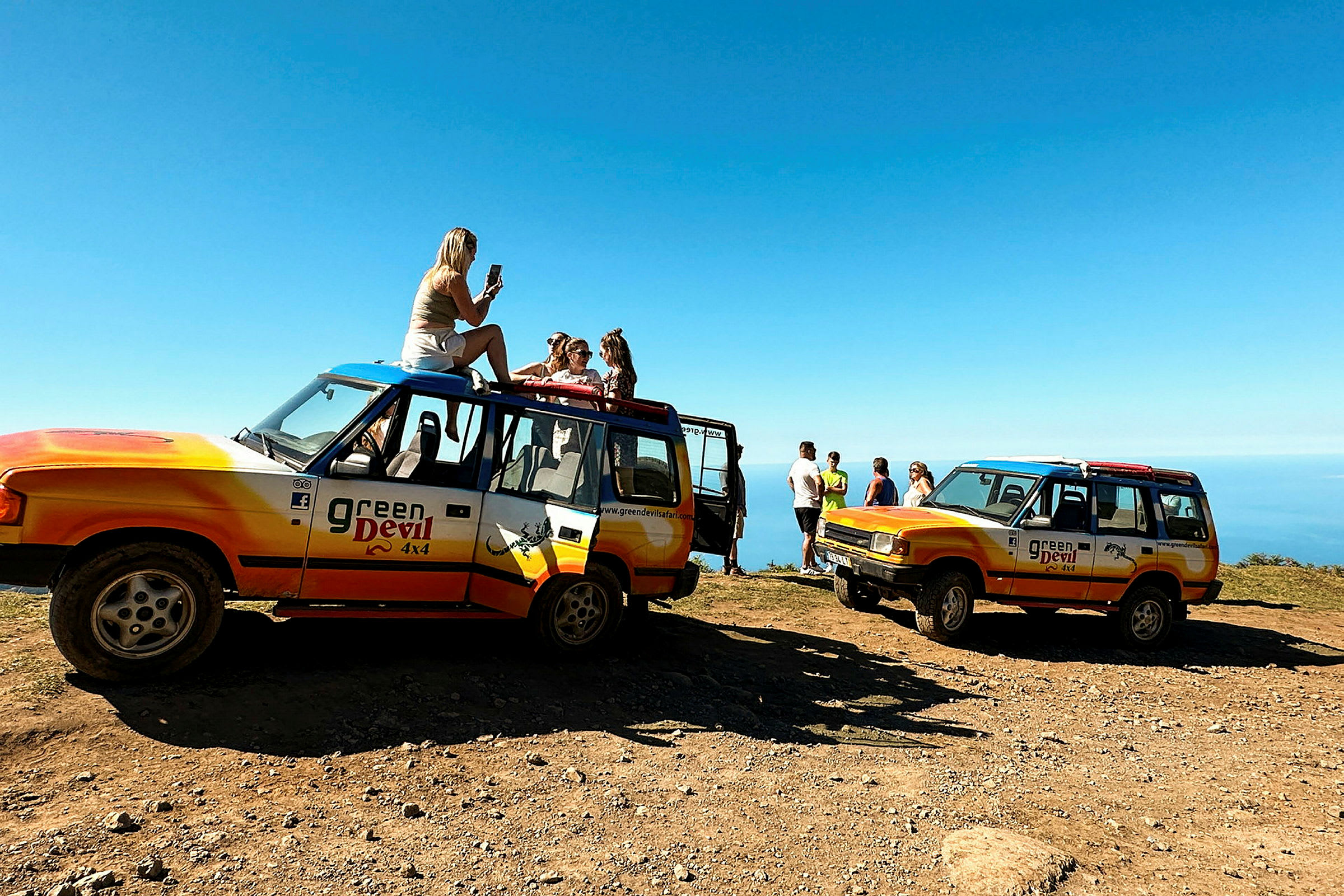 To terrengkjøretøy med "Green Devil 4x4"-logo er parkert på en grusvei med utsikt over havet. Folk står eller sitter nær kjøretøyene. (AI-generert tekst)