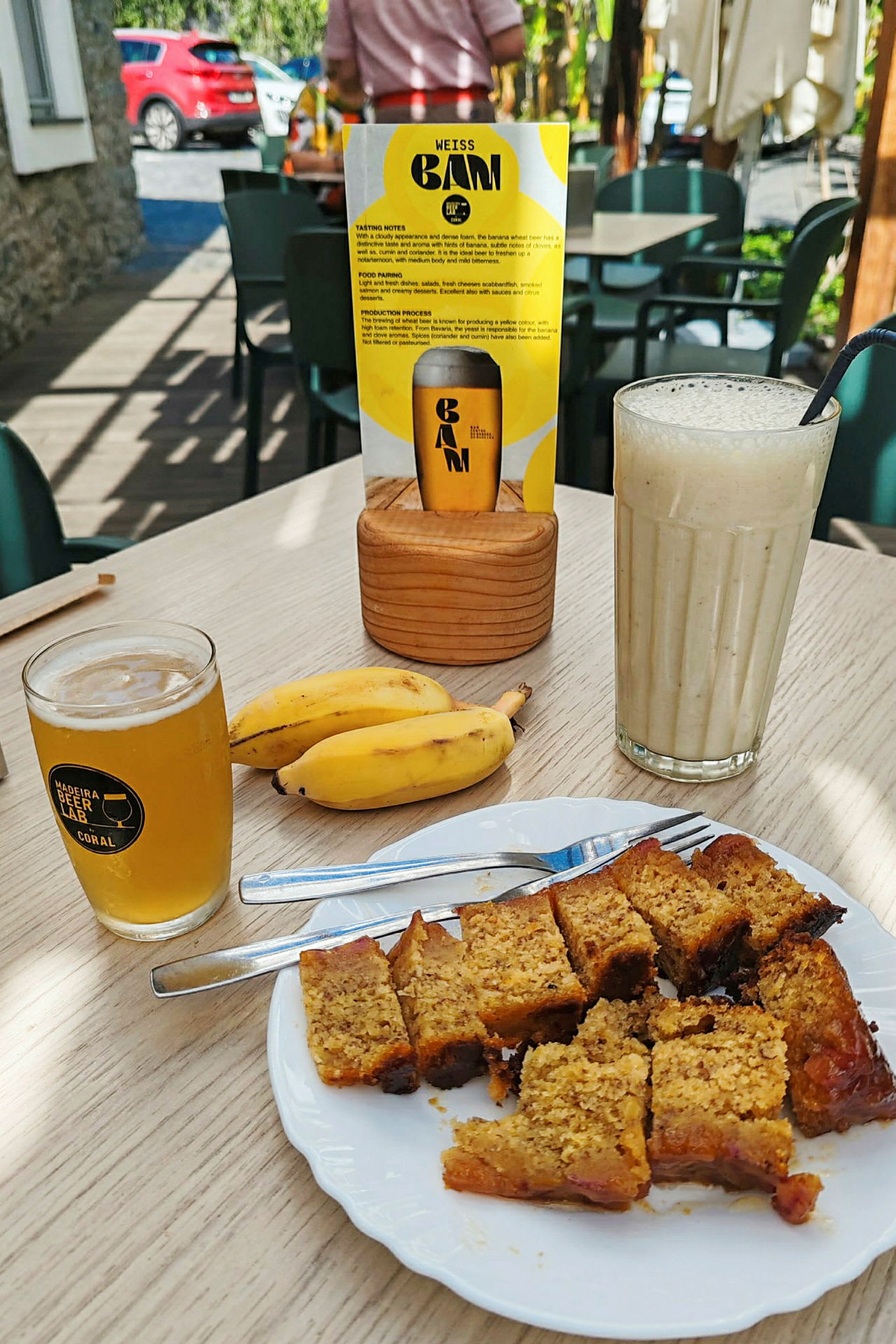En tallerken med kakestykker, to bananer, et glass øl og en milkshake på et bord. Bak sees en meny og sittemøbler utendørs. (AI-generert tekst)
