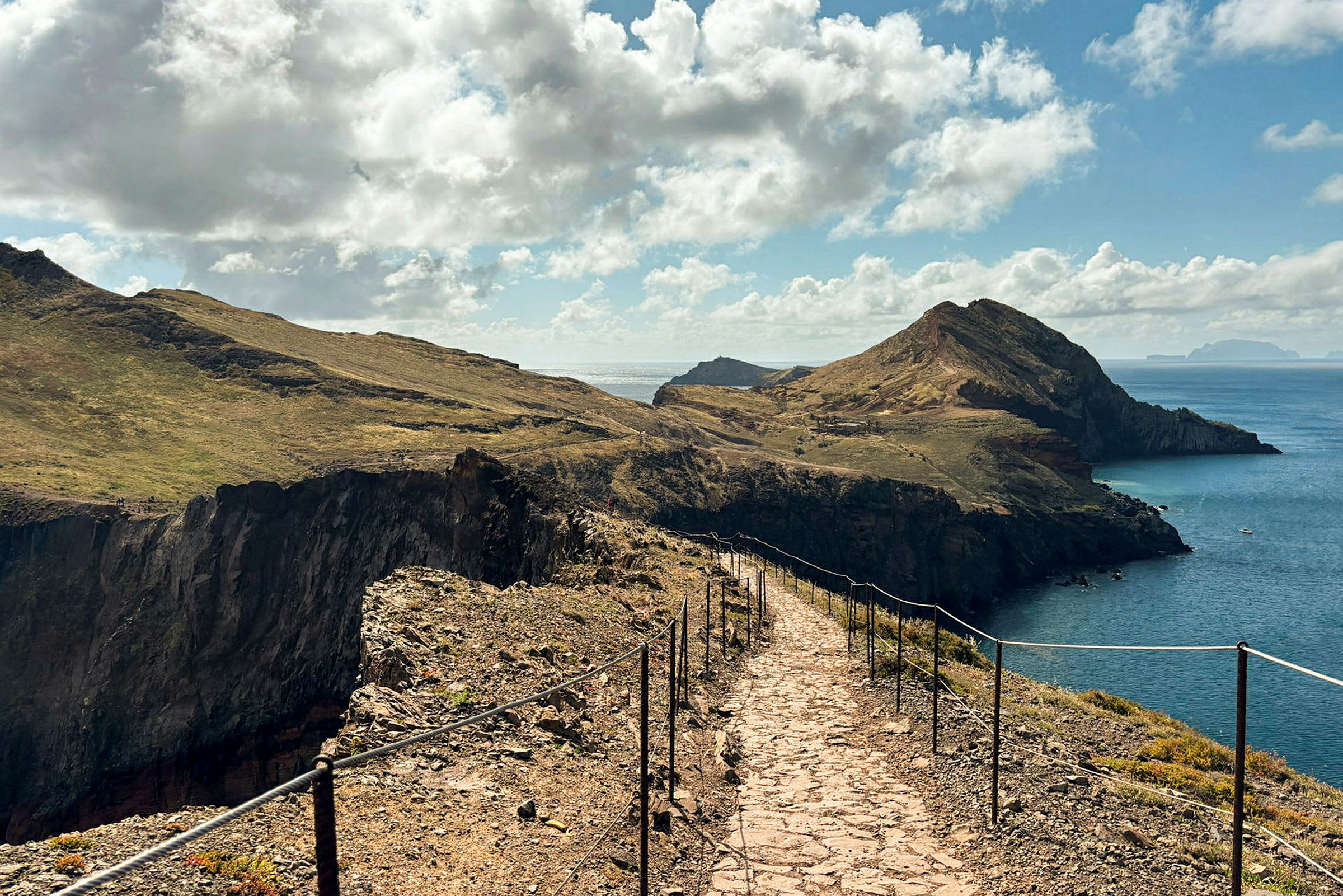 Et landskapsmotiv som viser en steinbelagt sti med rekkverk på begge sider. Stien går langs en klippe omgitt av vegetasjon og leder til fjell nær kysten med havet i bakgrunnen. (AI-generert tekst)