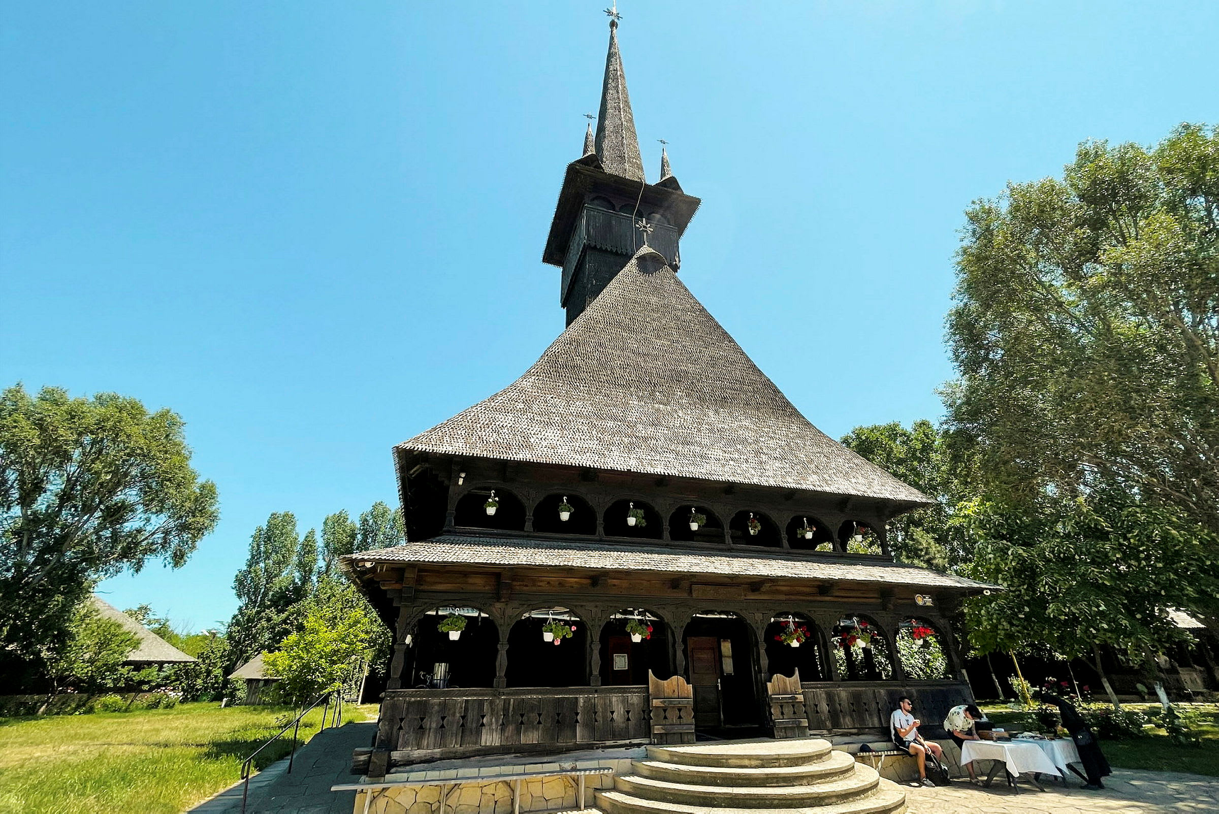 Nærbilde av en trekirke med spiss tak og dekorative elementer, omgitt av grønplanter og mennesker. (AI-generert tekst)